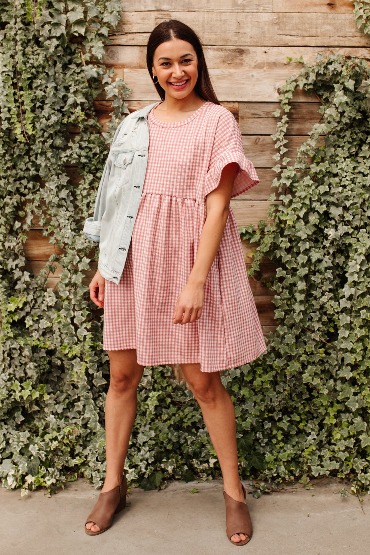 Smiling woman wearing pink gingham dress with flutter sleeves and brown open-toe booties standing by ivy-covered wooden wall