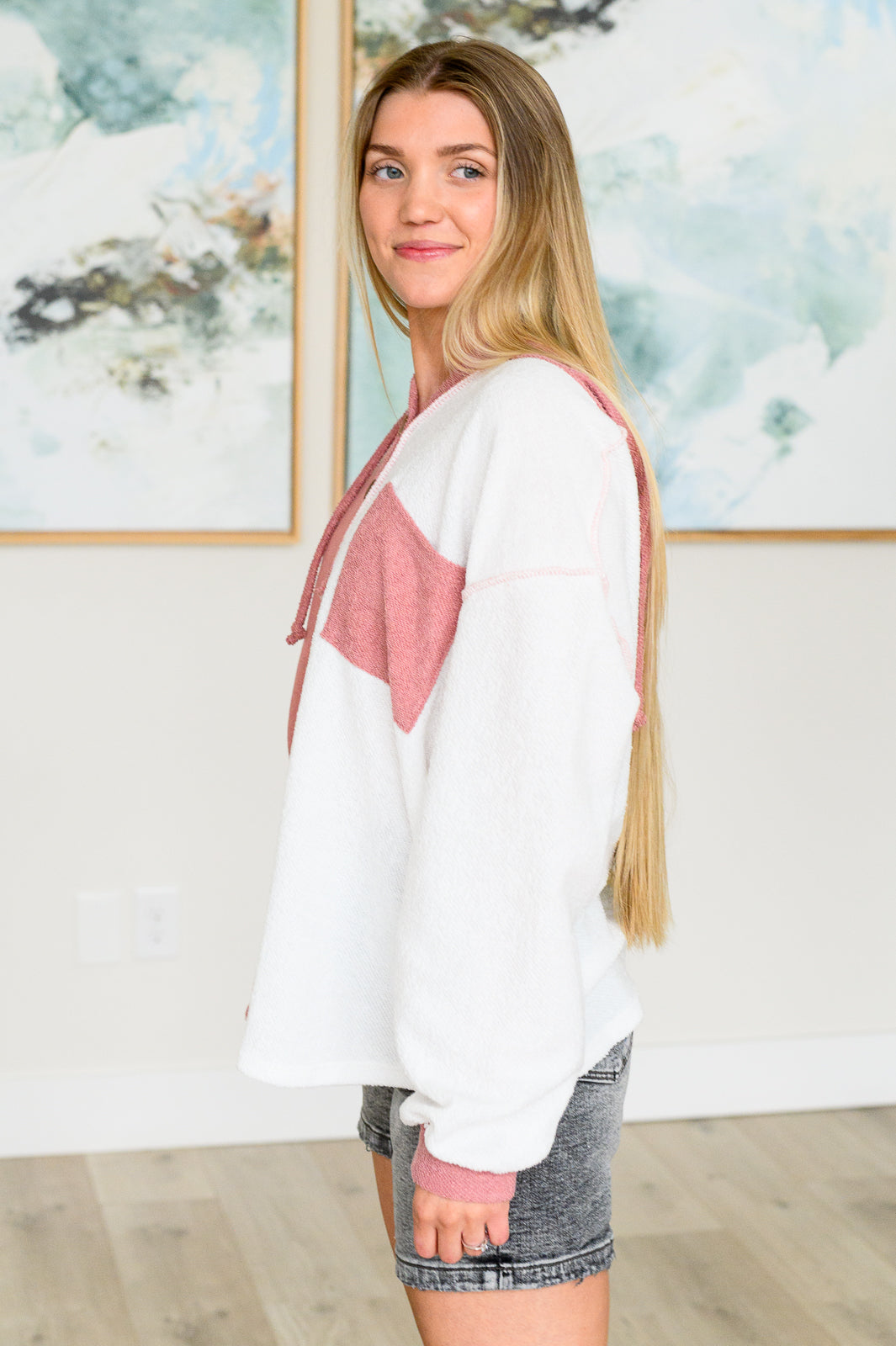 Young woman with long hair wearing a white and dusty rose color block hoodie standing indoors facing sideways