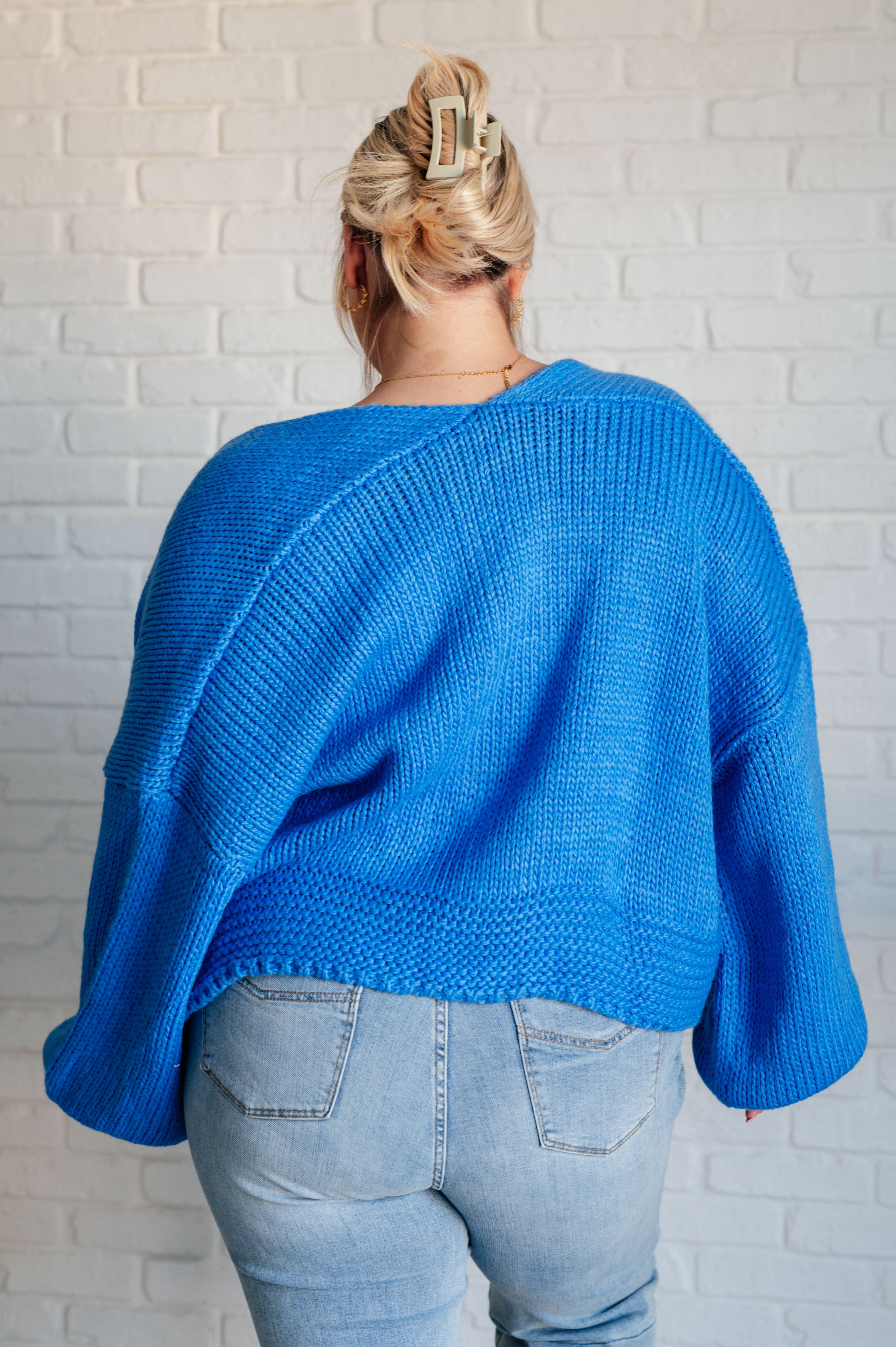 Back view of person wearing bright blue chunky knit cardigan paired with light wash jeans against white brick wall