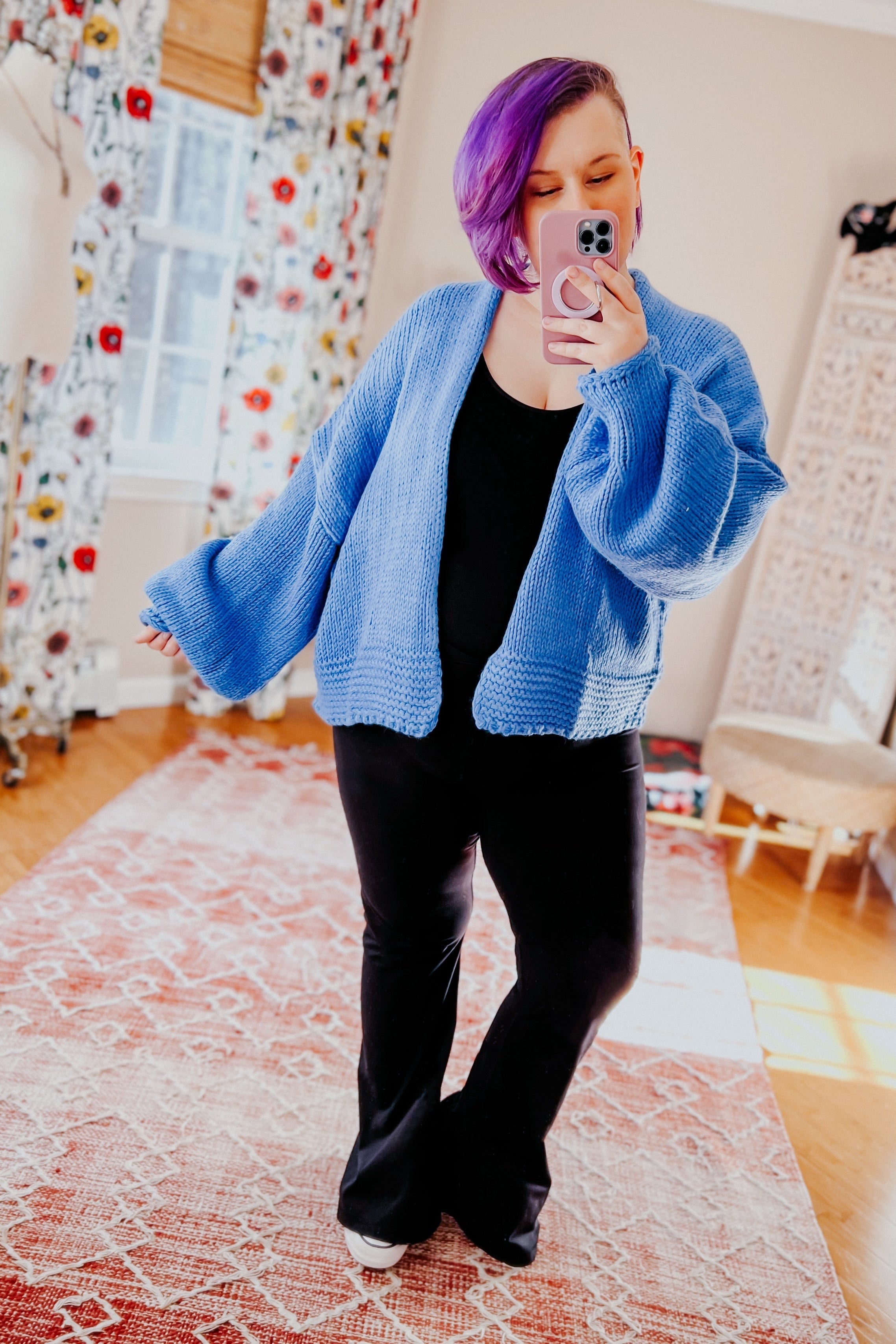 Person with purple hair wearing a light blue oversized cardigan over black outfit taking indoor mirror selfie in bright room