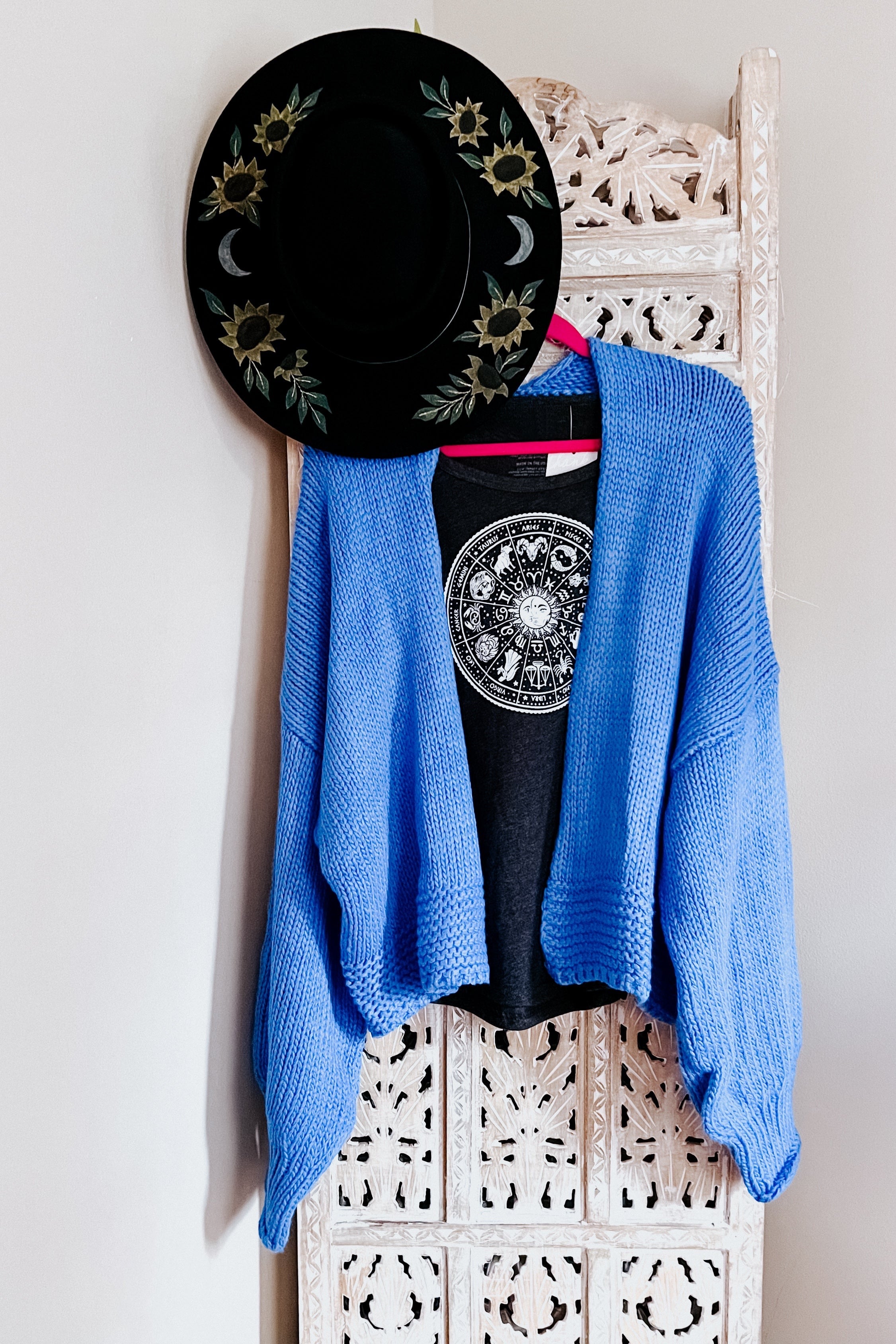 Bright blue loose knit cardigan layered over black zodiac graphic tee hanging on carved white screen with black floral hat