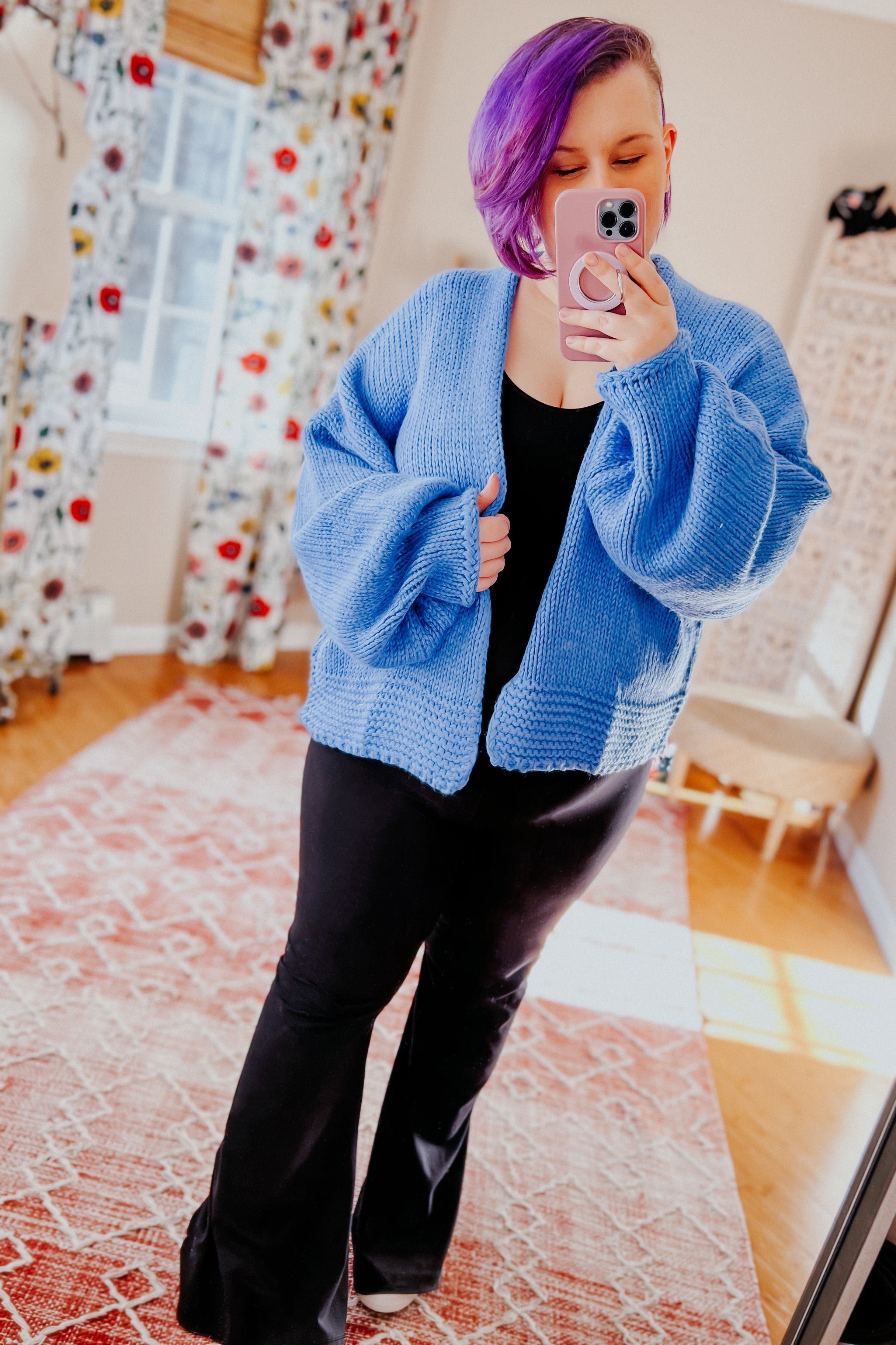 Woman with purple hair wearing light blue chunky knit cardigan over black top and pants taking selfie indoors