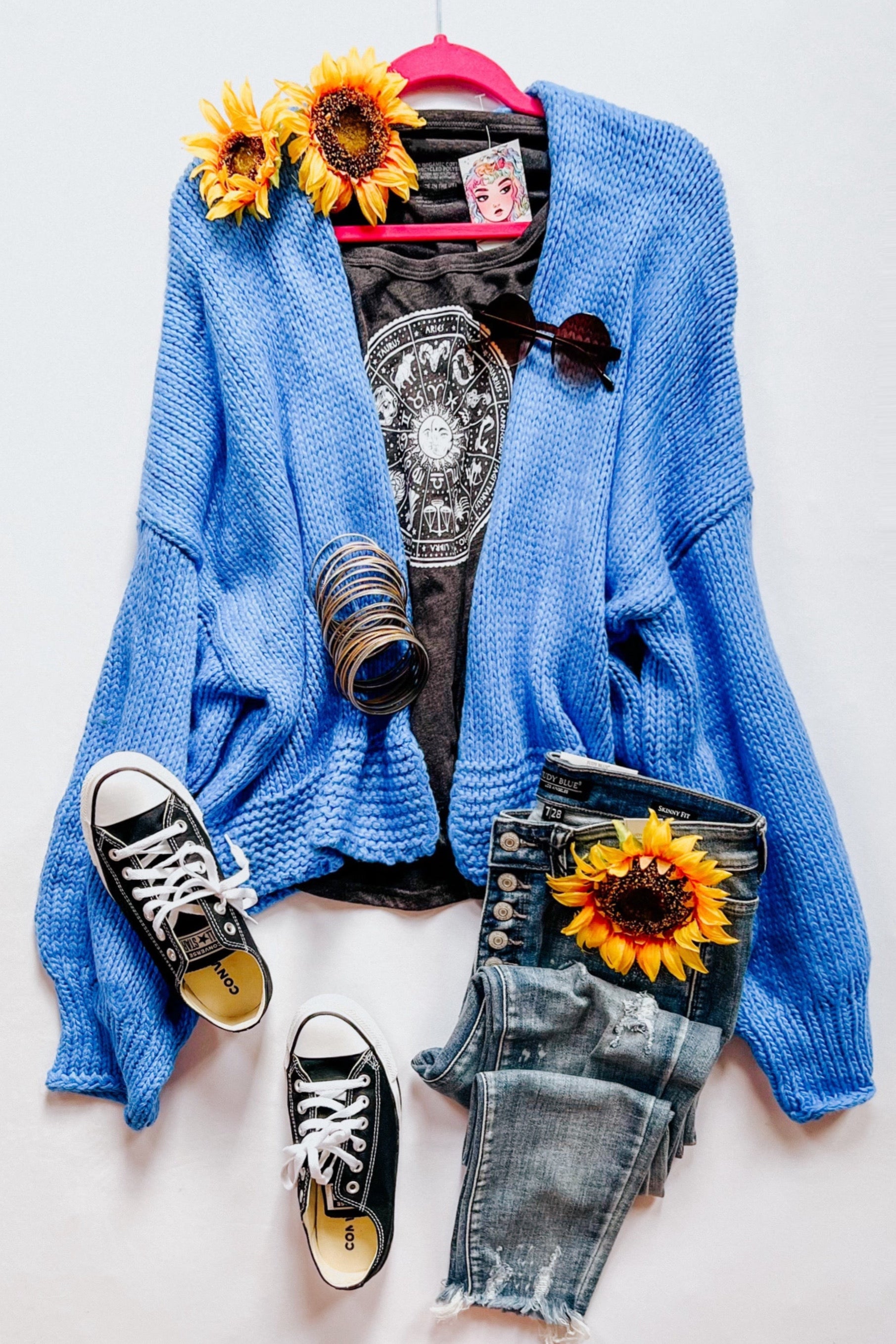Blue chunky knit cardigan styled with graphic tee, distressed jeans, black sneakers, sunglasses, bangles, and sunflowers on white background