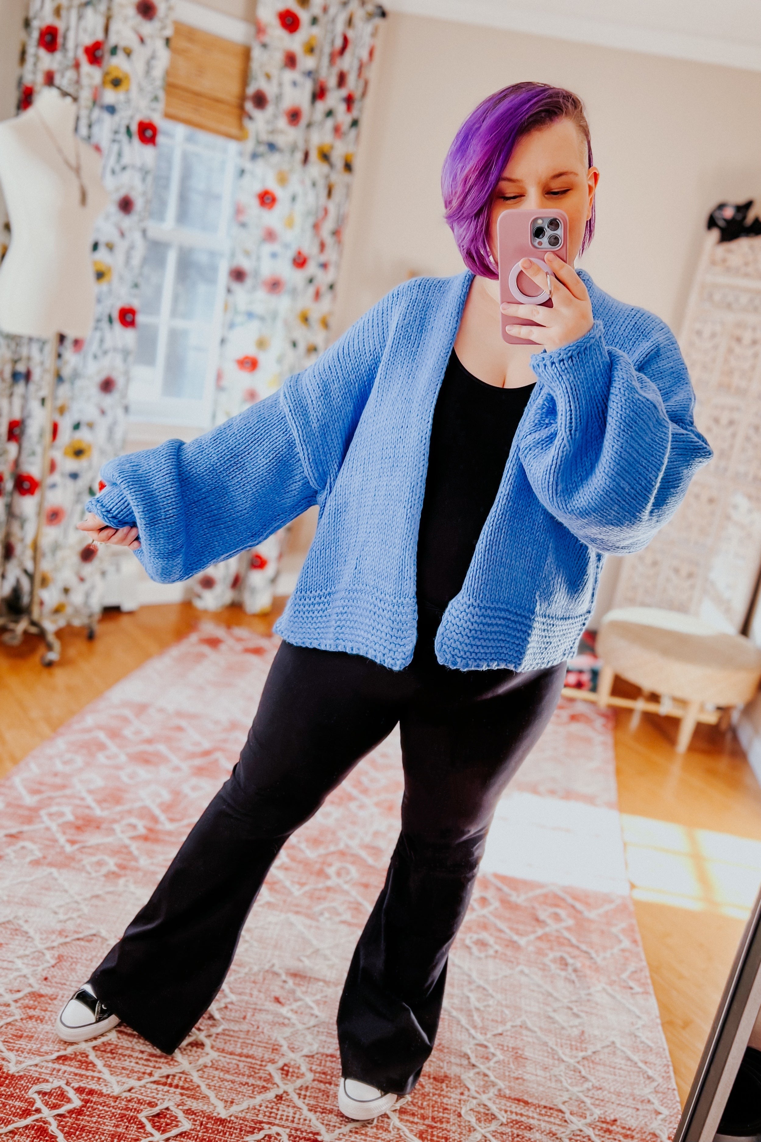 Woman with purple hair wearing a loose light blue cardigan over black outfit taking indoor mirror selfie