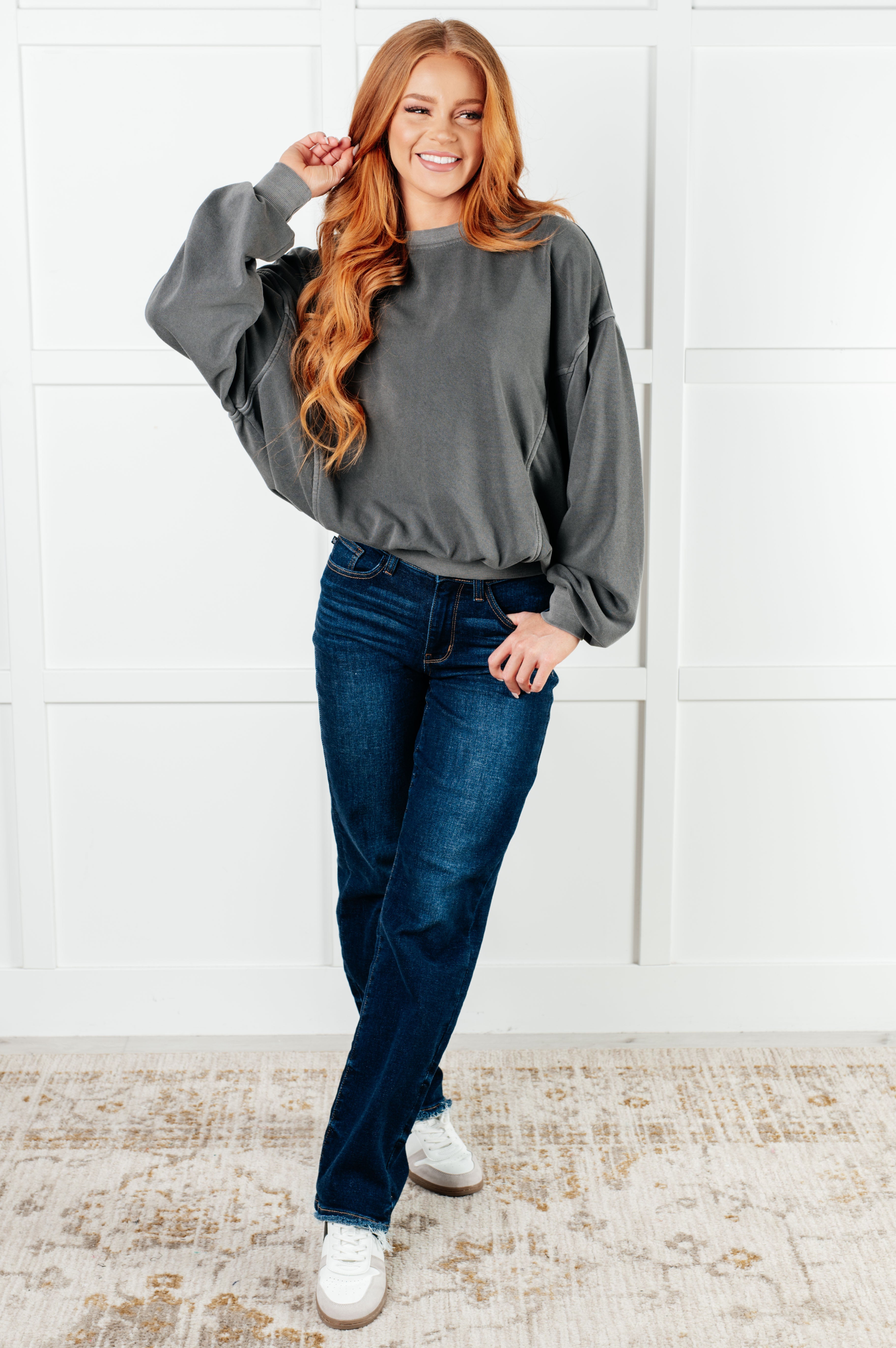 Smiling woman with long red hair wearing loose ash black pullover and dark blue jeans standing on patterned rug