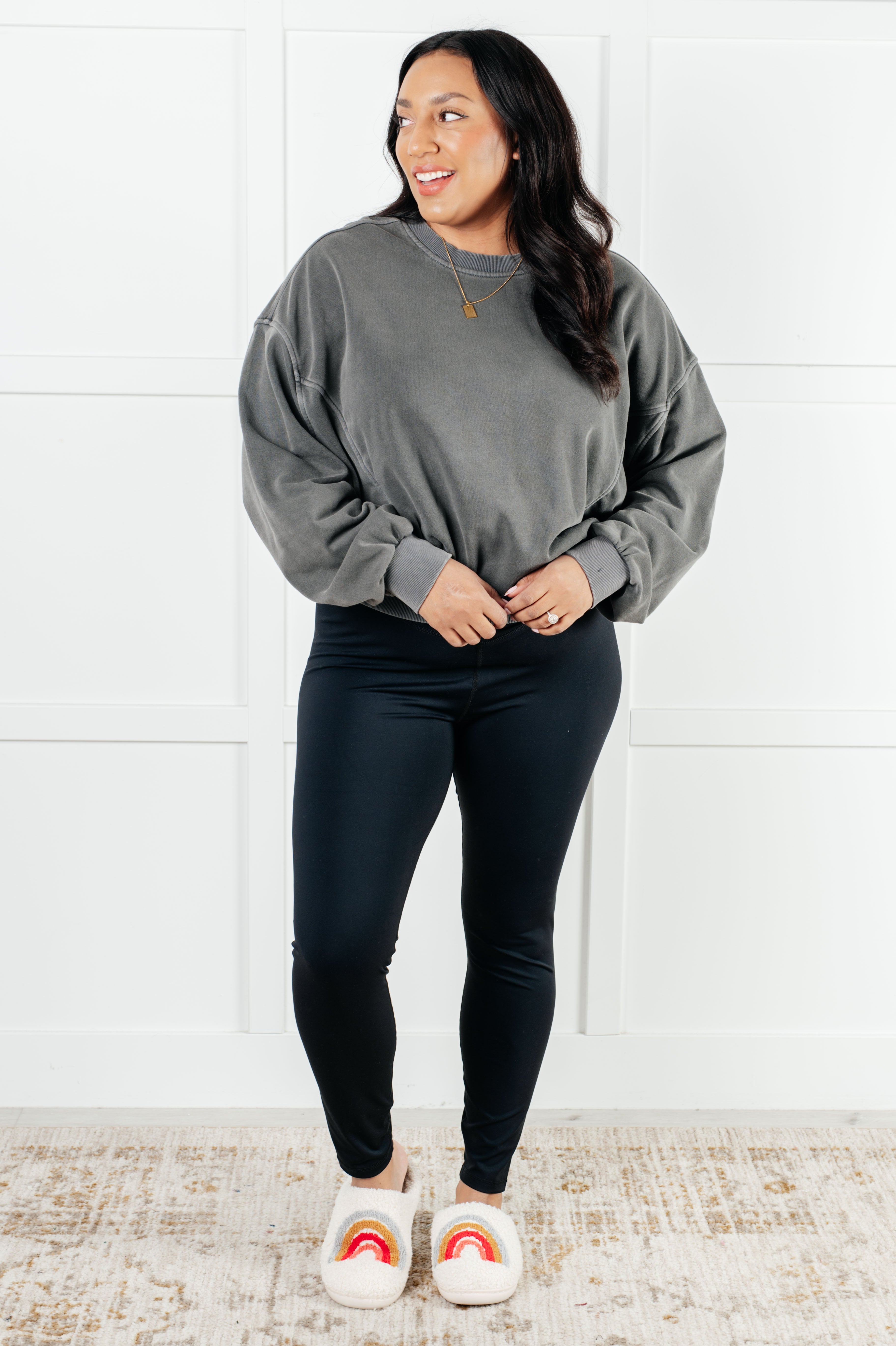 Woman wearing loose ash black pullover and black fitted leggings standing on patterned rug against white paneled wall