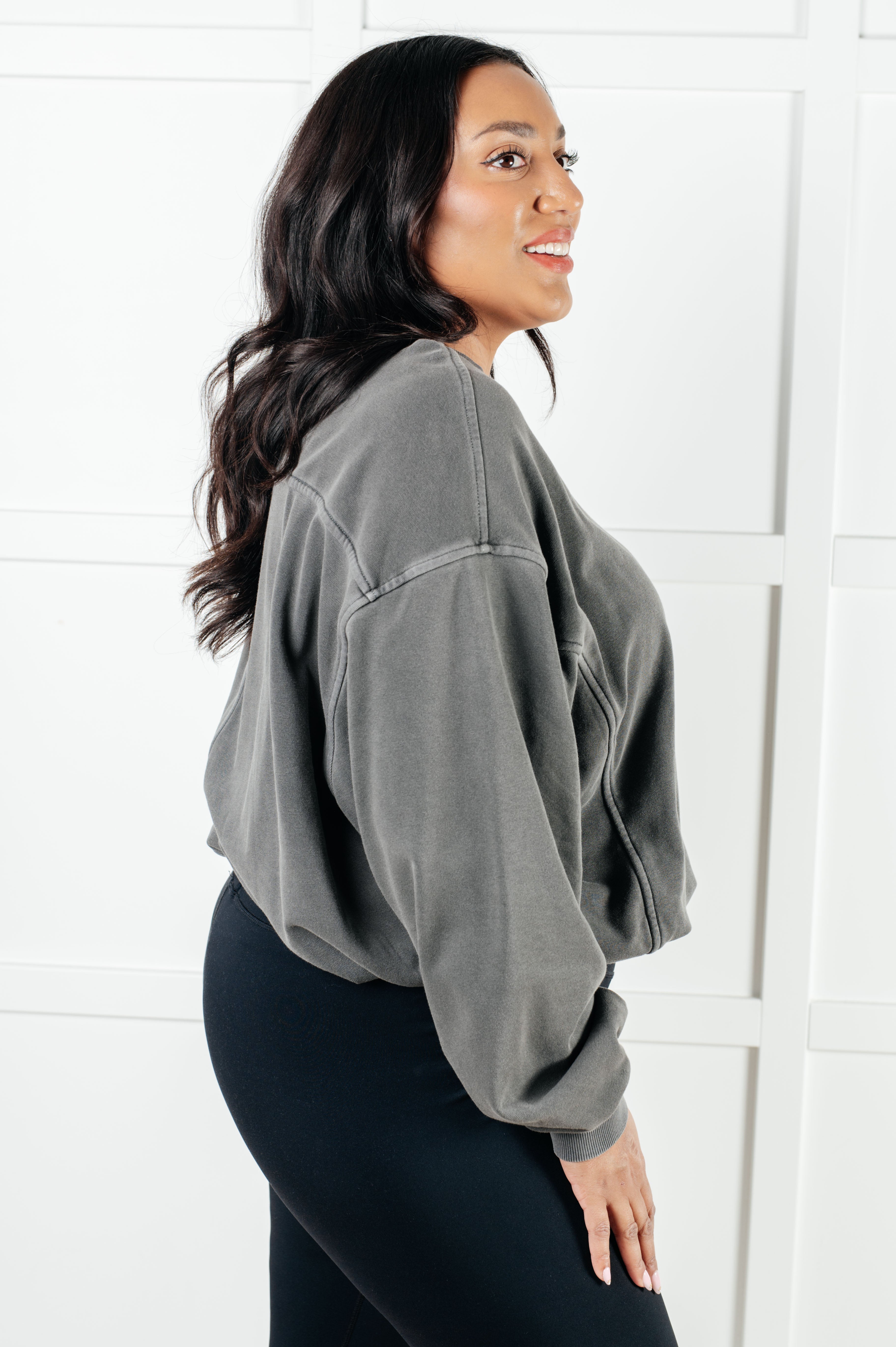 Side profile of woman wearing ash black relaxed pullover and black leggings against white paneled wall
