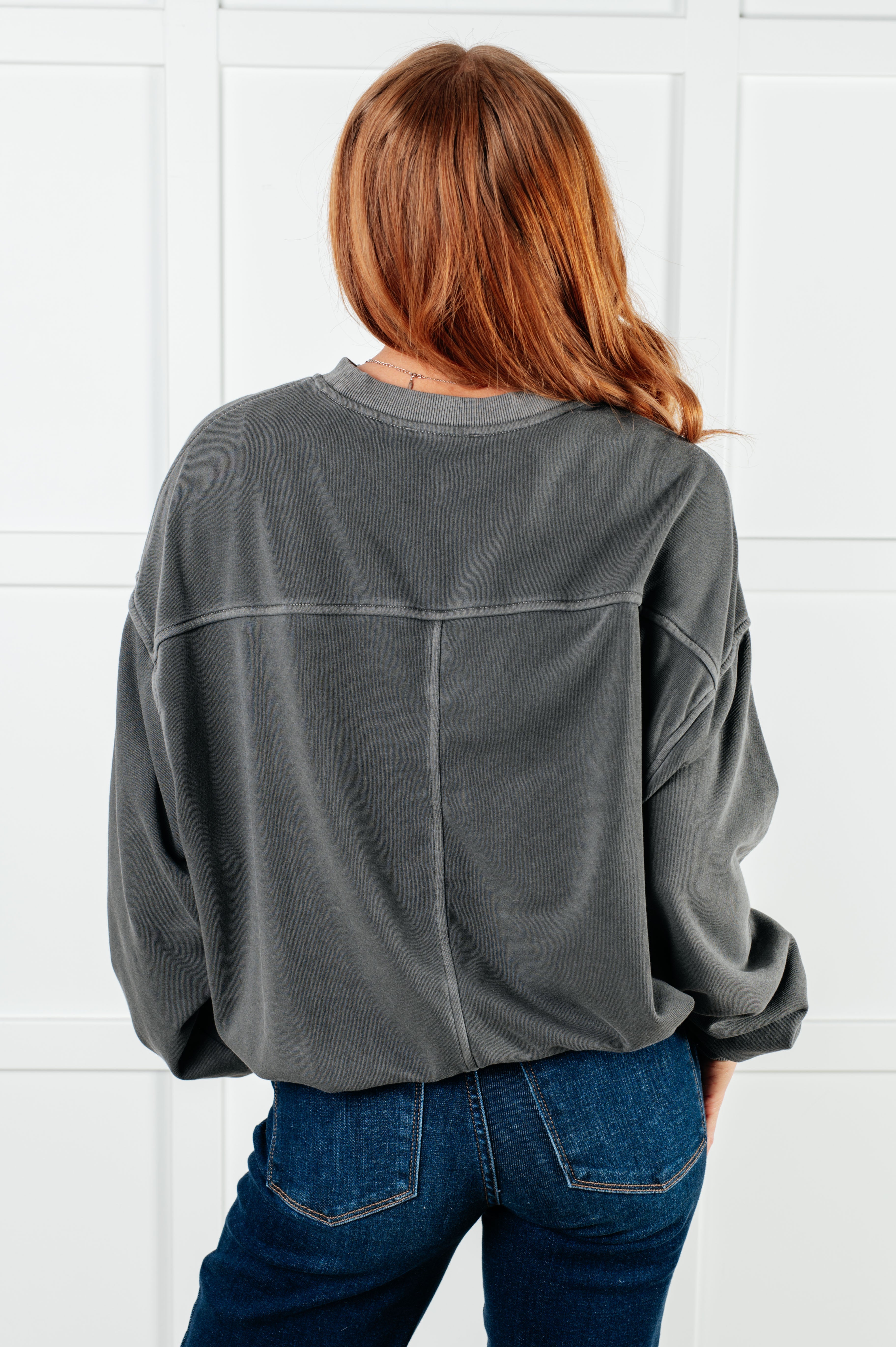 Back view of person with auburn hair wearing loose ash black pullover and dark blue jeans against white background