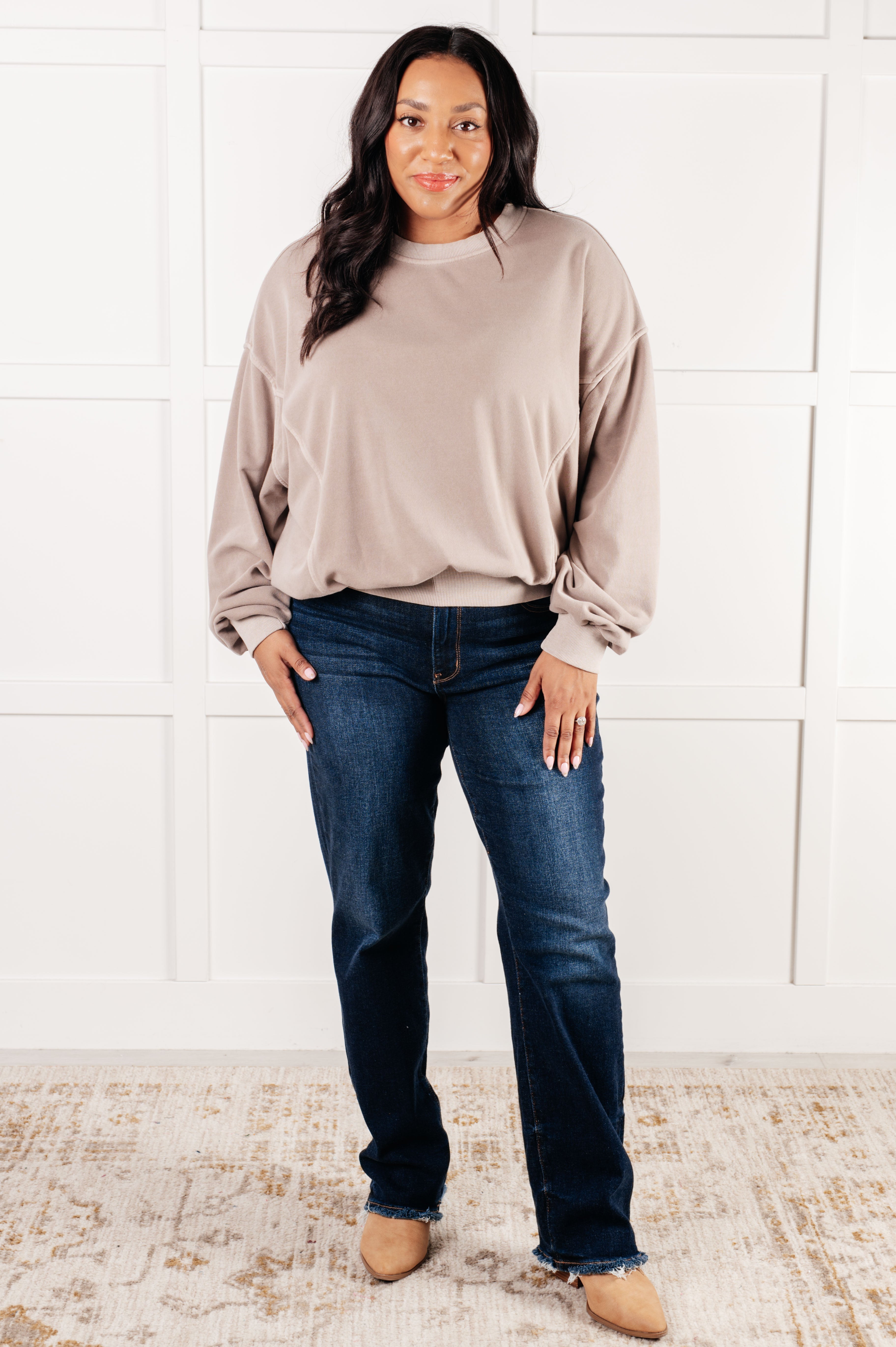 Woman with long dark hair wearing loose ash mocha pullover and dark blue jeans standing on patterned rug against white wall
