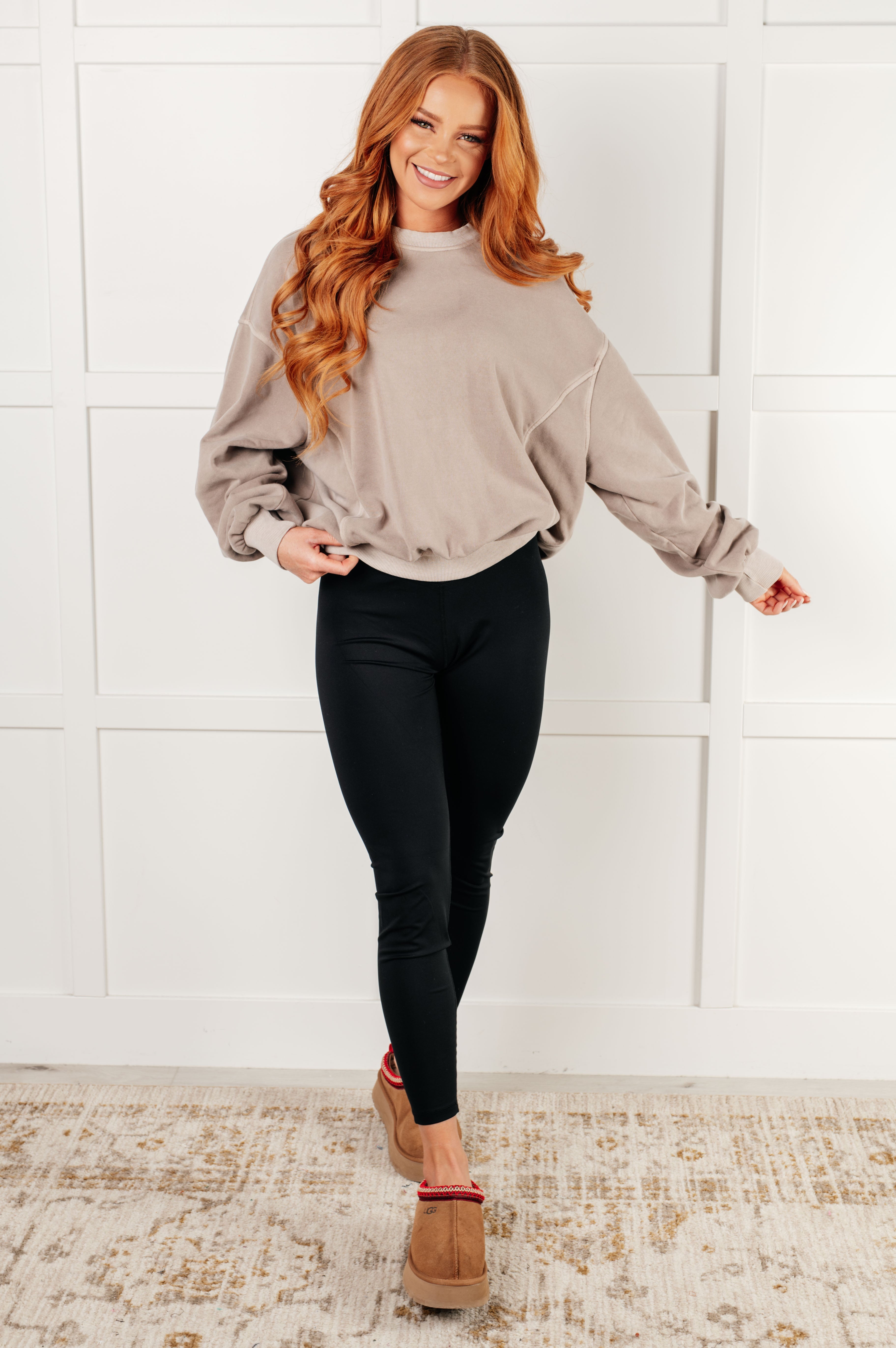 Smiling woman with long red hair wearing loose ash mocha pullover and black leggings standing on rug against white wall