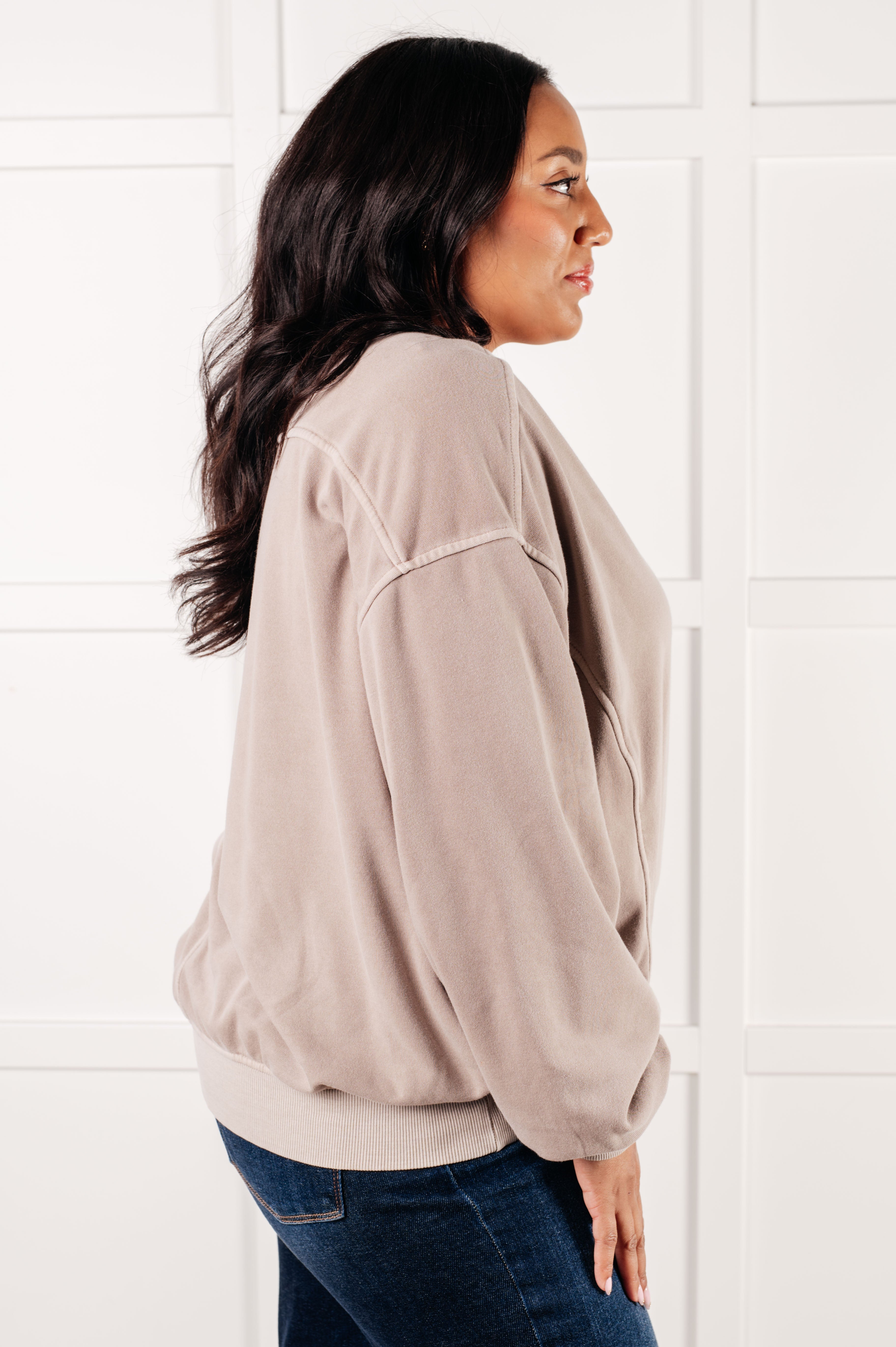 Profile of woman with dark wavy hair wearing oversized ash mocha pullover and blue jeans against white paneled wall