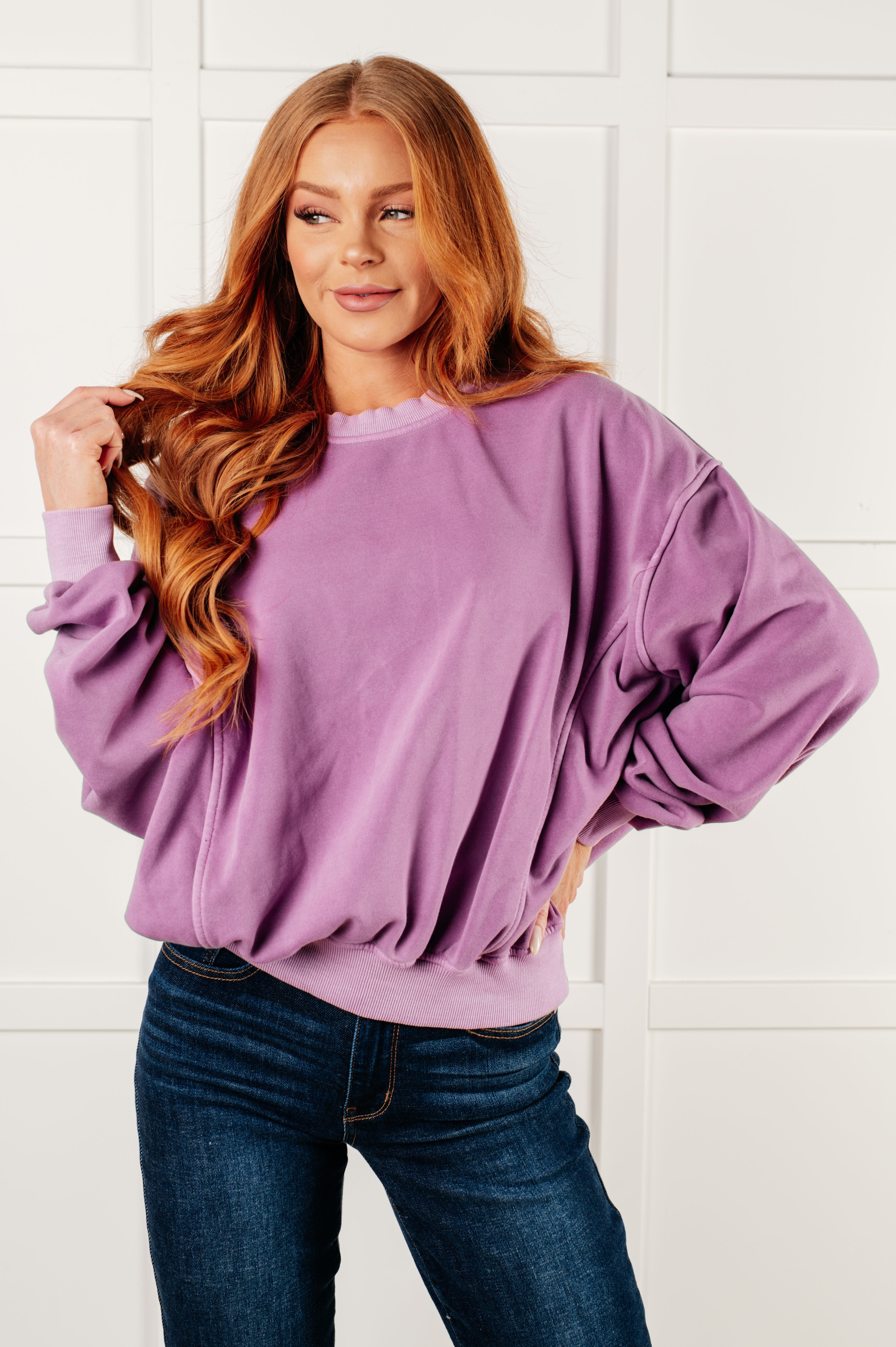Woman with long red wavy hair wearing loose violet pullover and dark blue jeans standing against white paneled wall