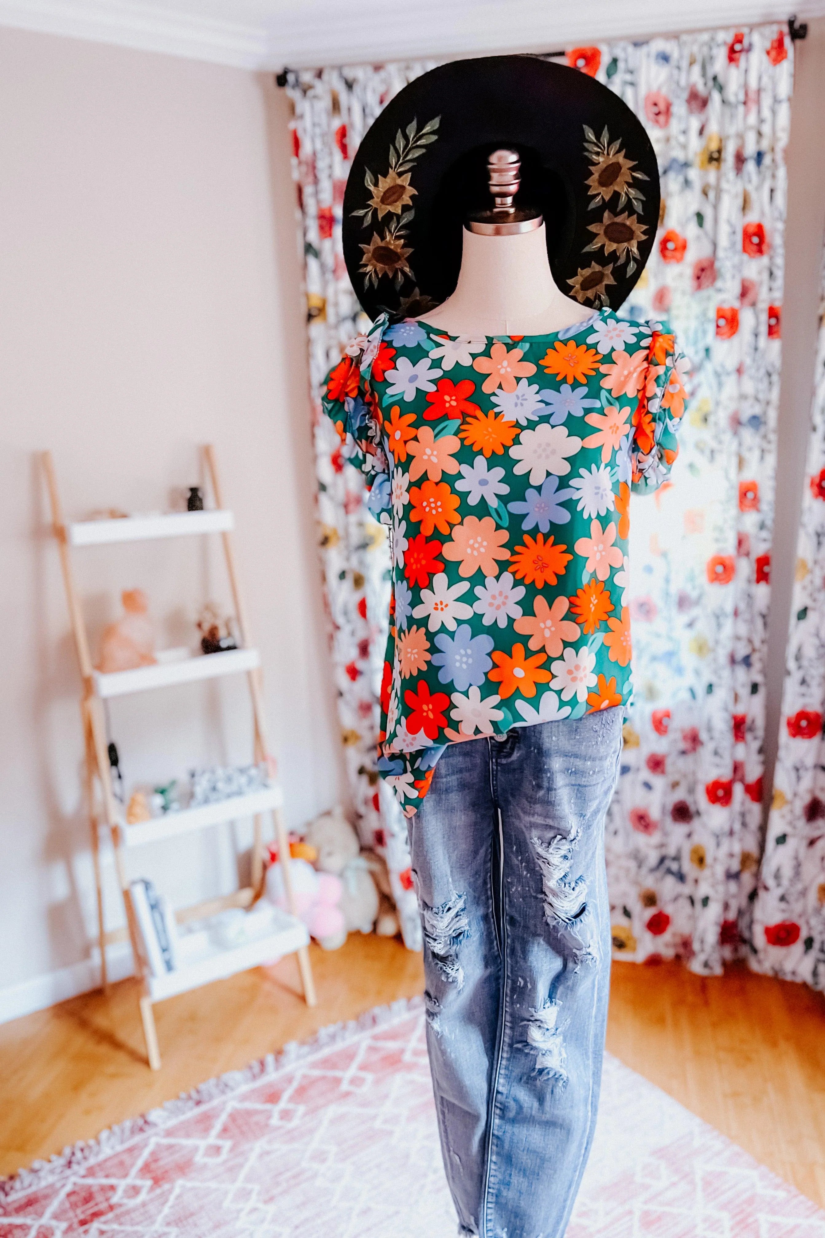 Mannequin wearing vibrant floral double ruffle sleeve top paired with distressed denim jeans and black sunflower hat indoors