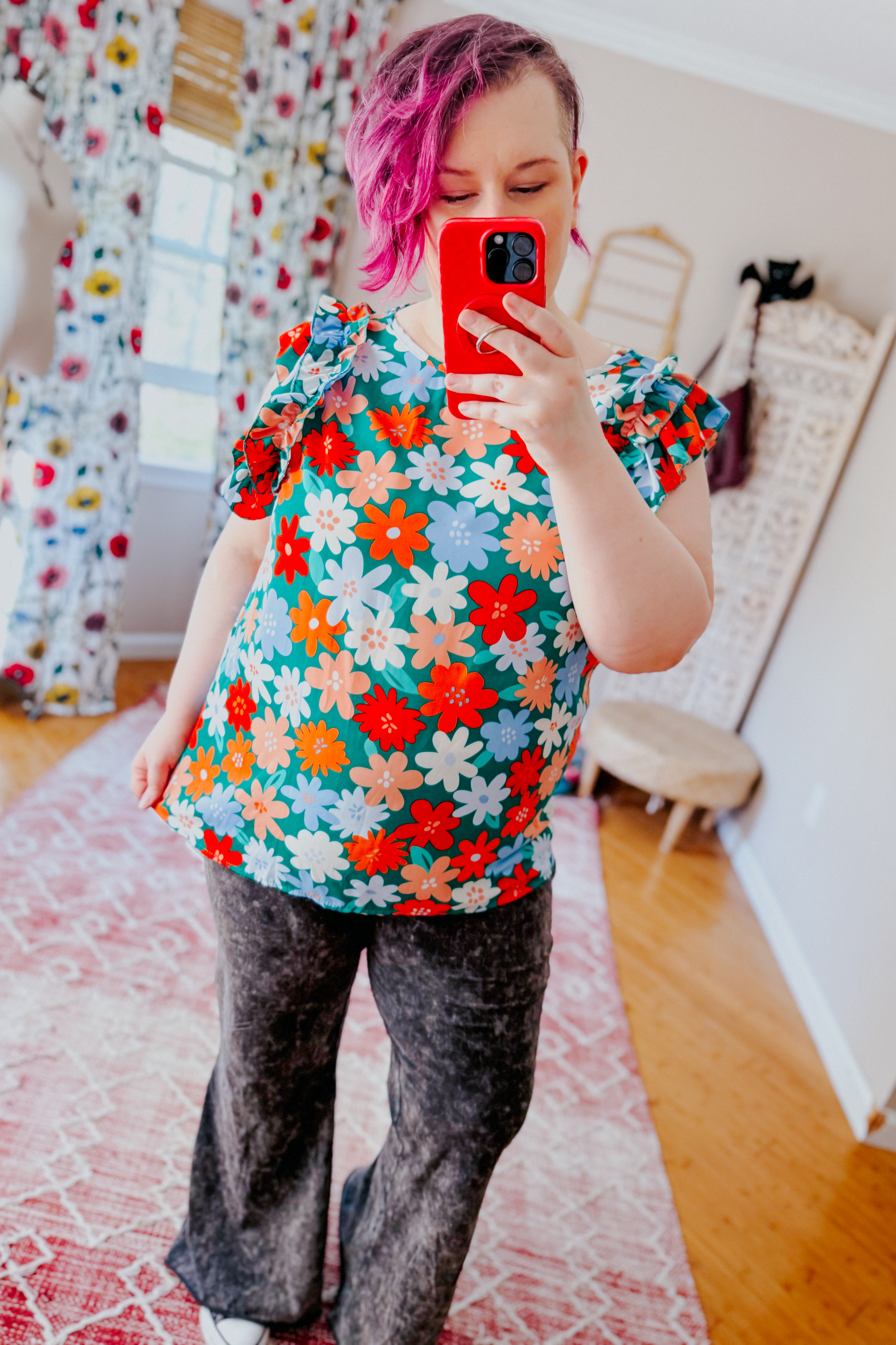 Woman with pink hair wearing colorful floral double ruffle sleeve top and acid wash black flare pants taking mirror selfie in bright room