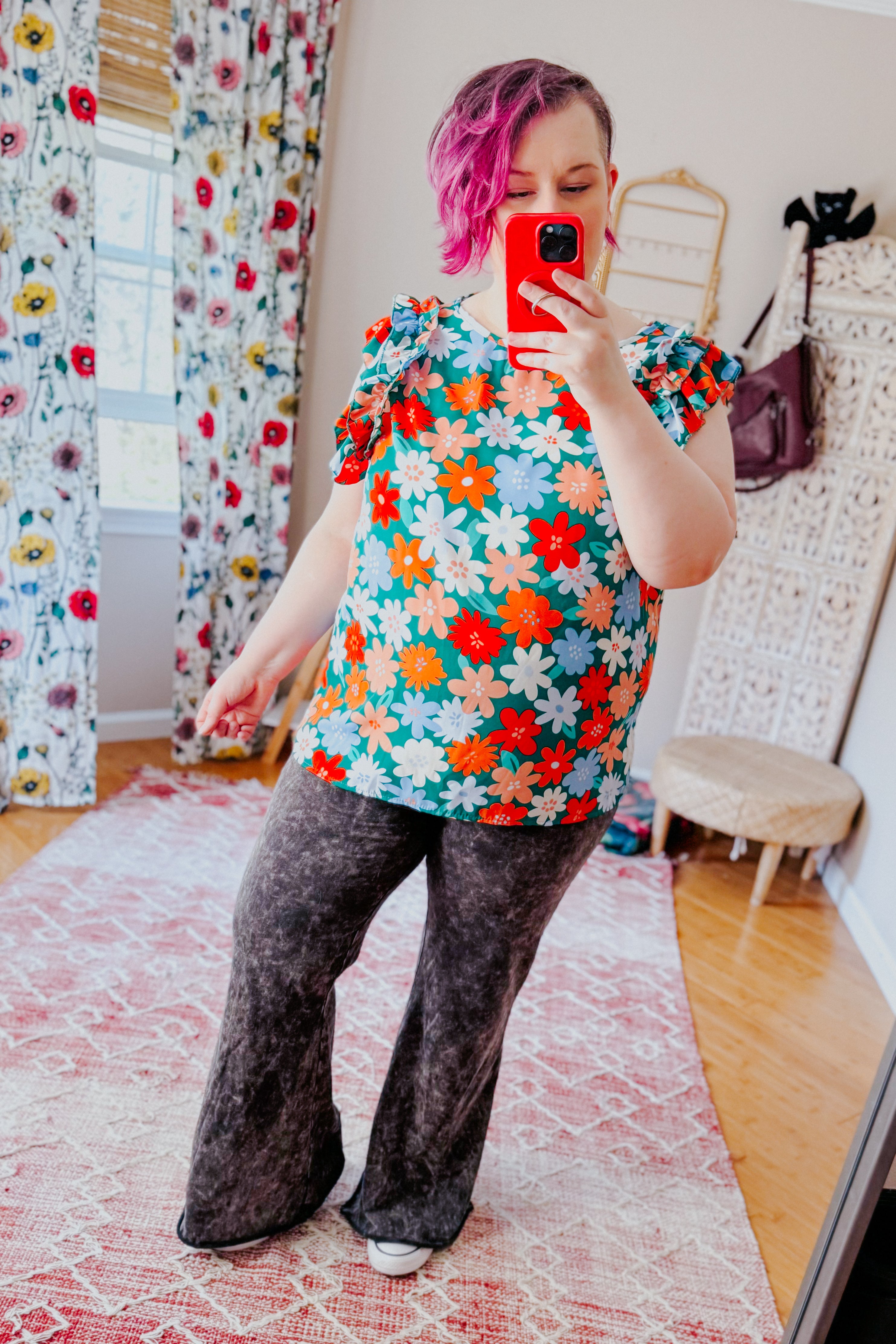 Woman with short pink hair wearing colorful floral double ruffle sleeve top and black washed flare pants taking mirror selfie in room