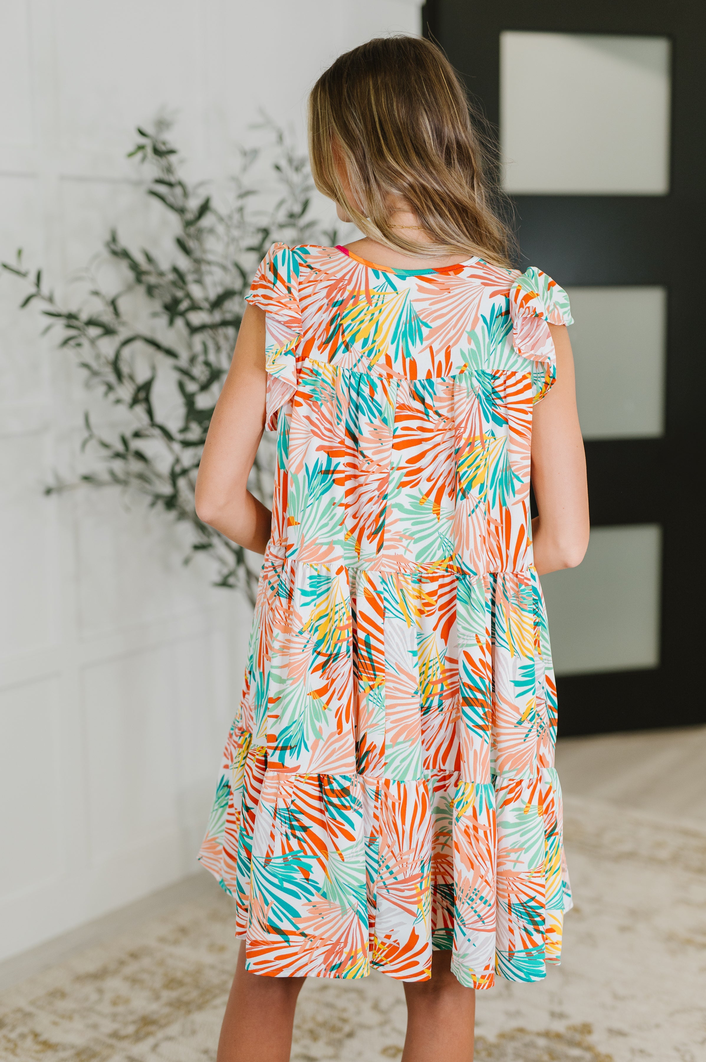Back view of girl wearing Ivory and orange patterned tiered dress with flutter sleeves in a bright indoor space