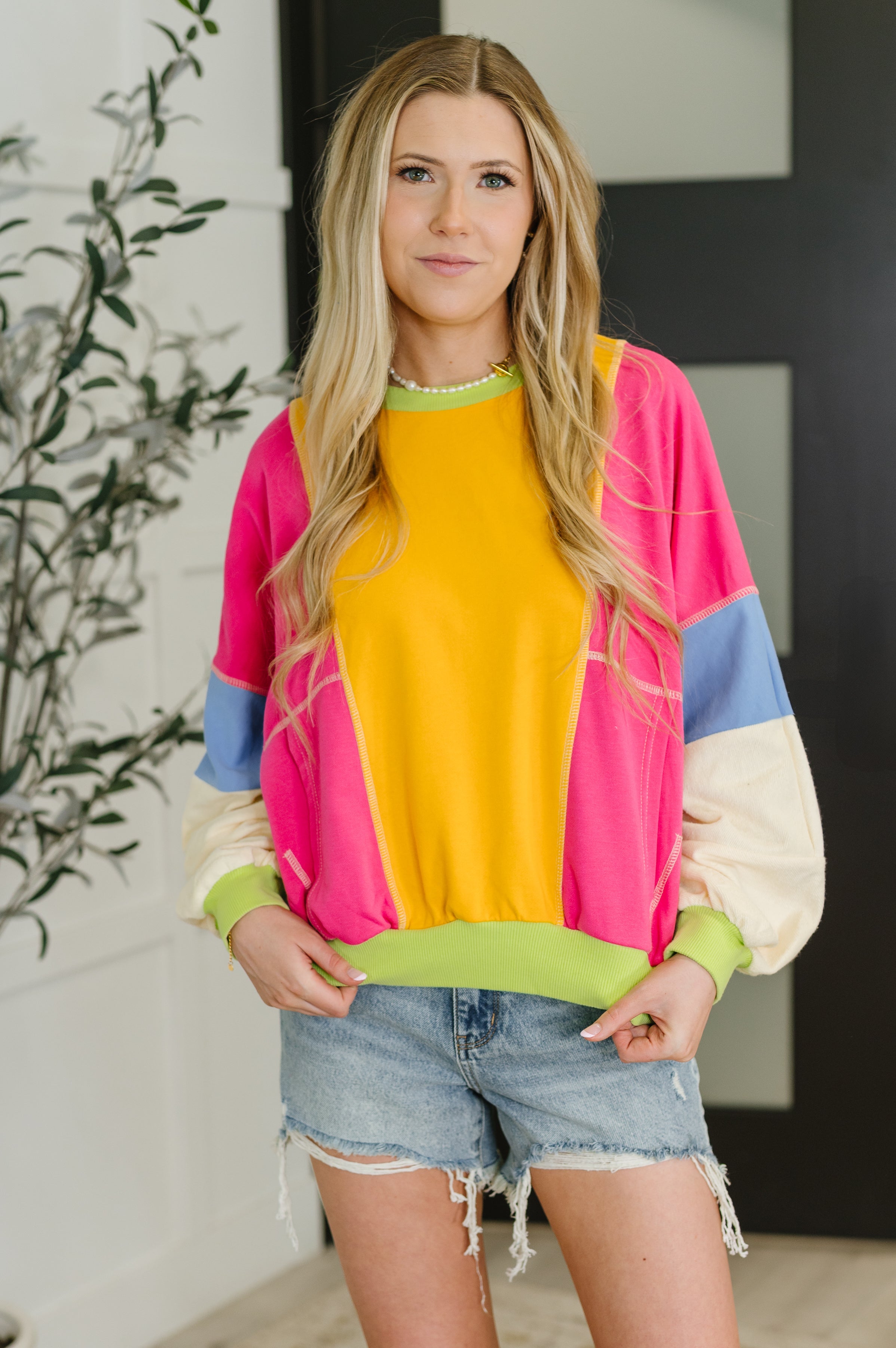 Woman wearing a yellow and fuchsia color block pullover with blue and cream sleeves and denim shorts indoors