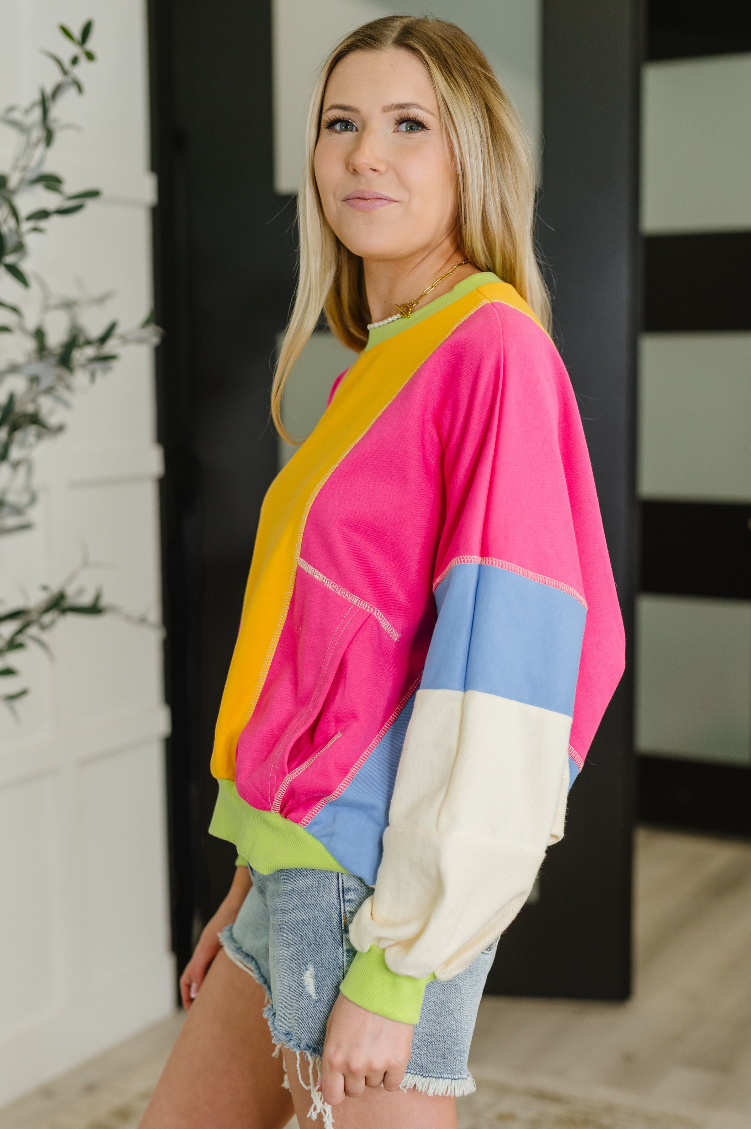 Woman in a bright yellow and fuchsia color block pullover with blue and cream sleeves and denim shorts indoors