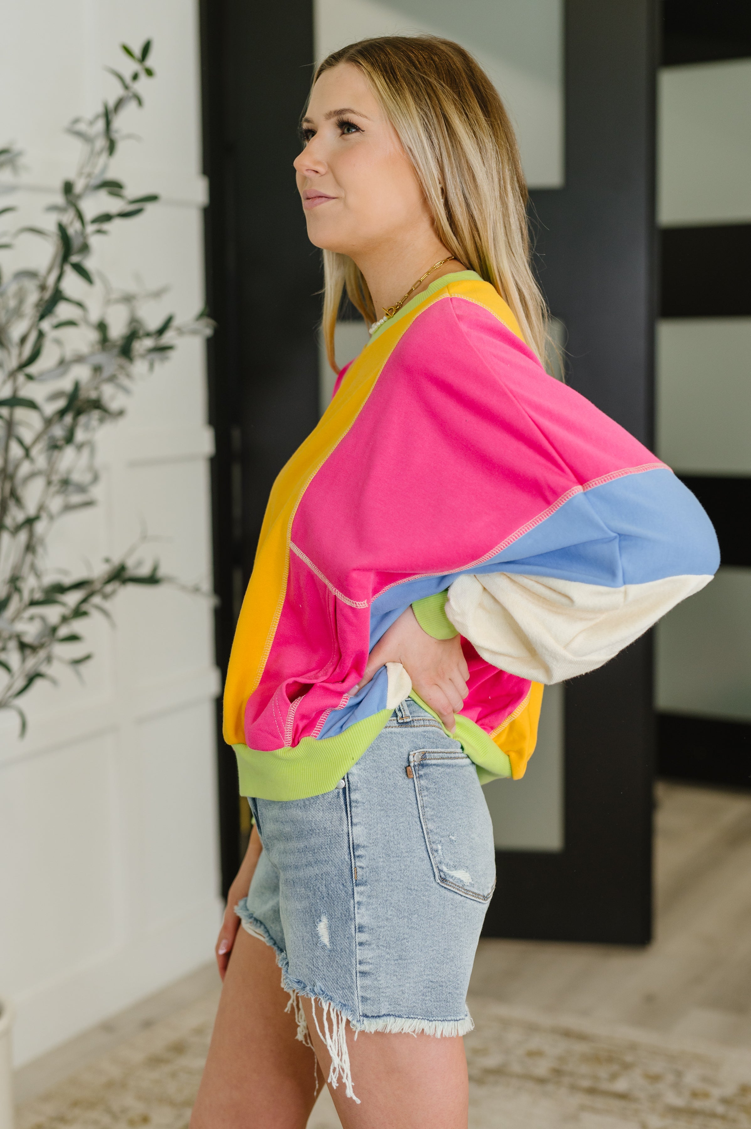 Side view of a woman in a yellow and fuchsia color block pullover with blue and cream dolman sleeves and denim shorts indoors