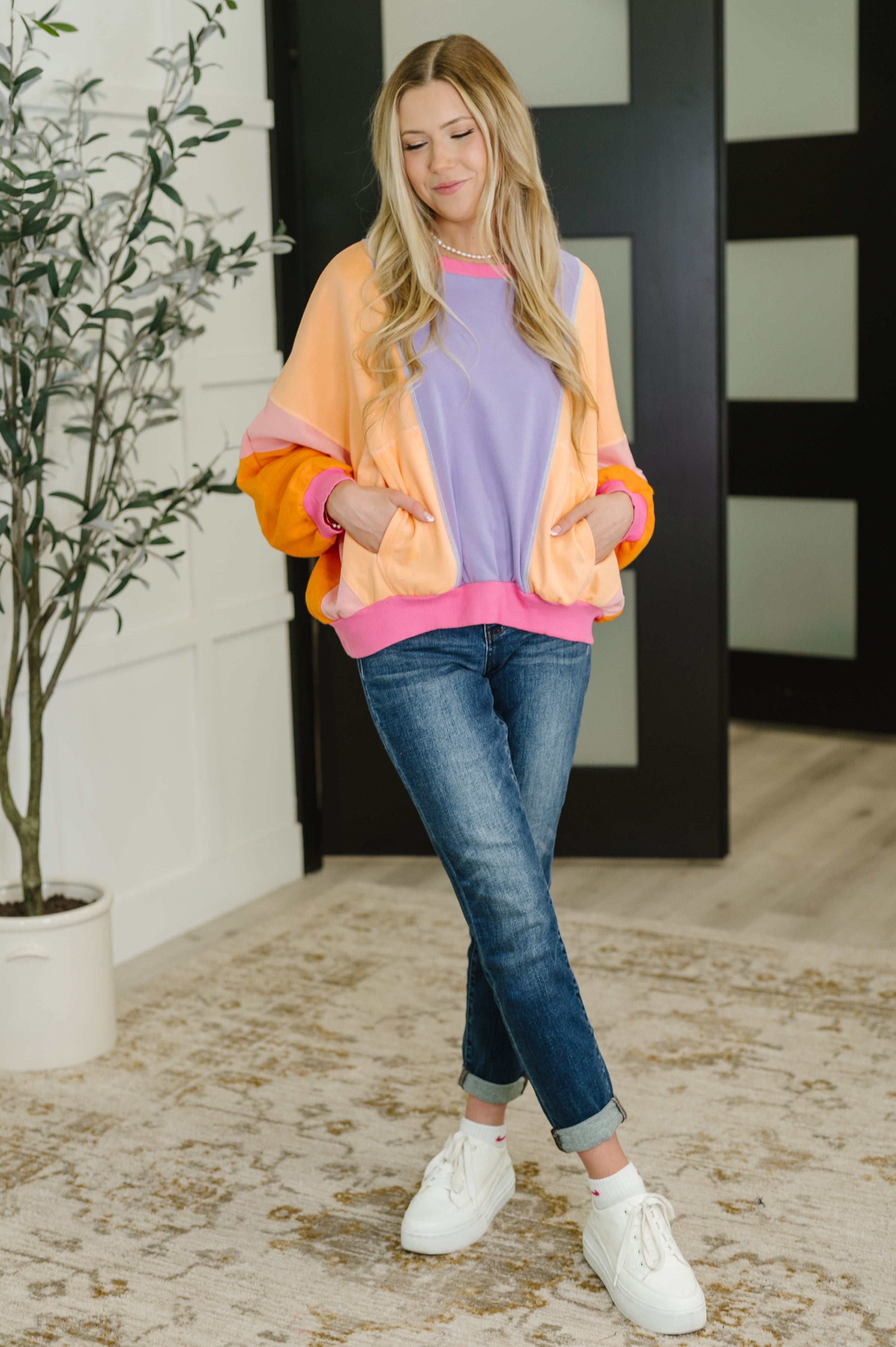 Woman modeling a purple and yellow color block pullover with peach sleeves and pink trim over jeans indoors