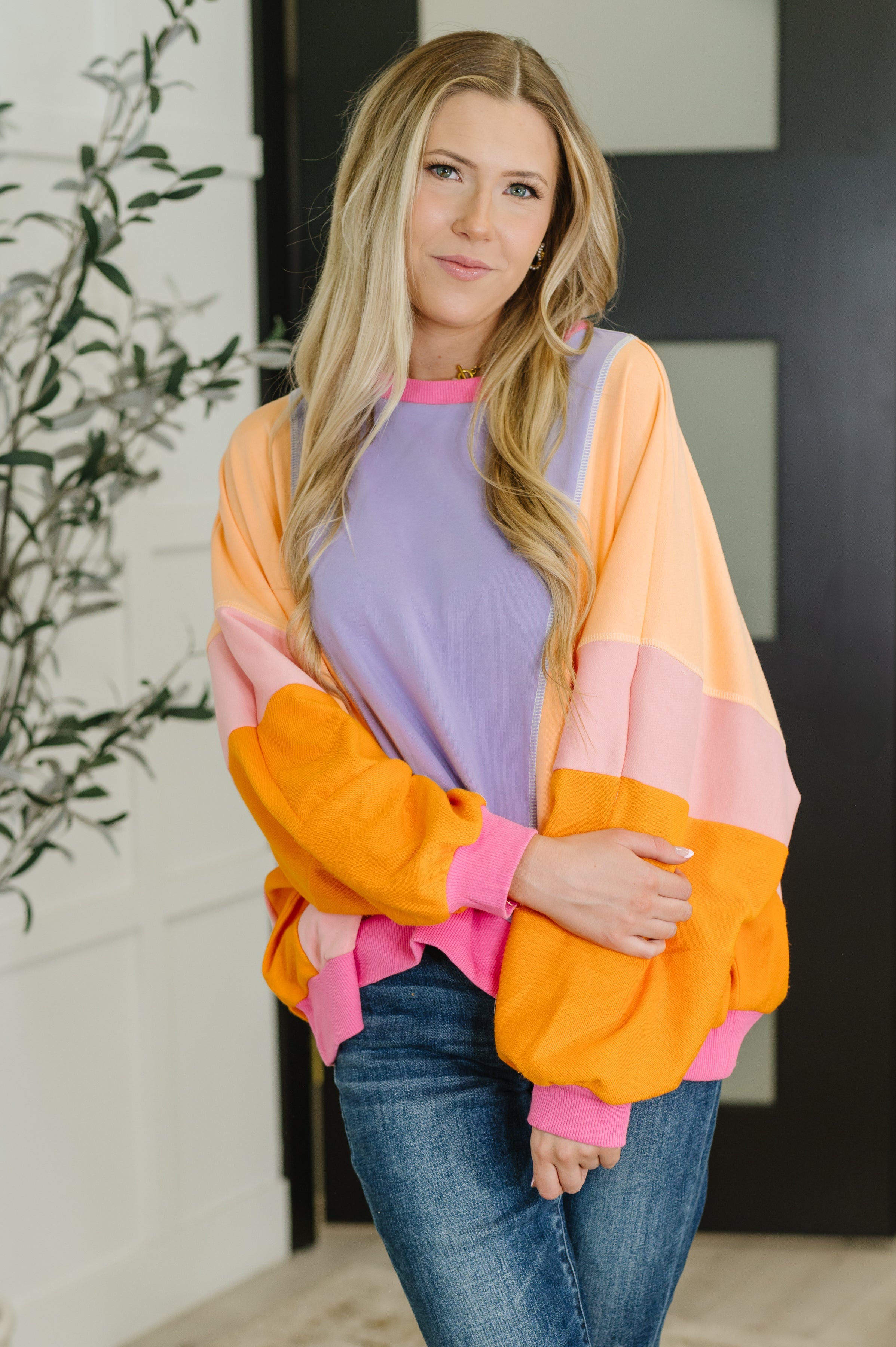 Woman wearing a purple and yellow color block pullover with orange and pink sleeves over jeans indoors