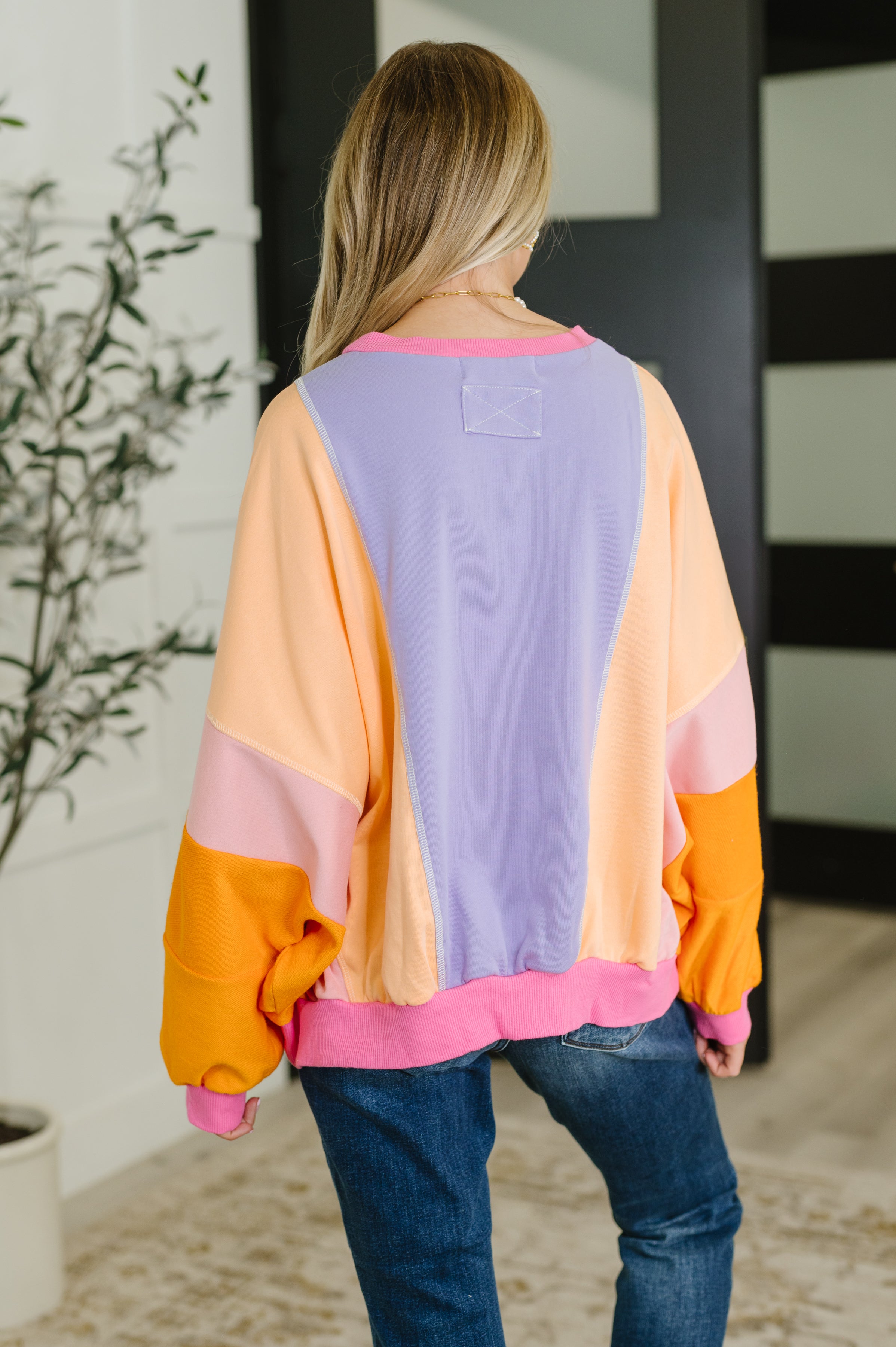 Back view of woman wearing a purple and yellow color block pullover with pink and orange sleeves over jeans indoors