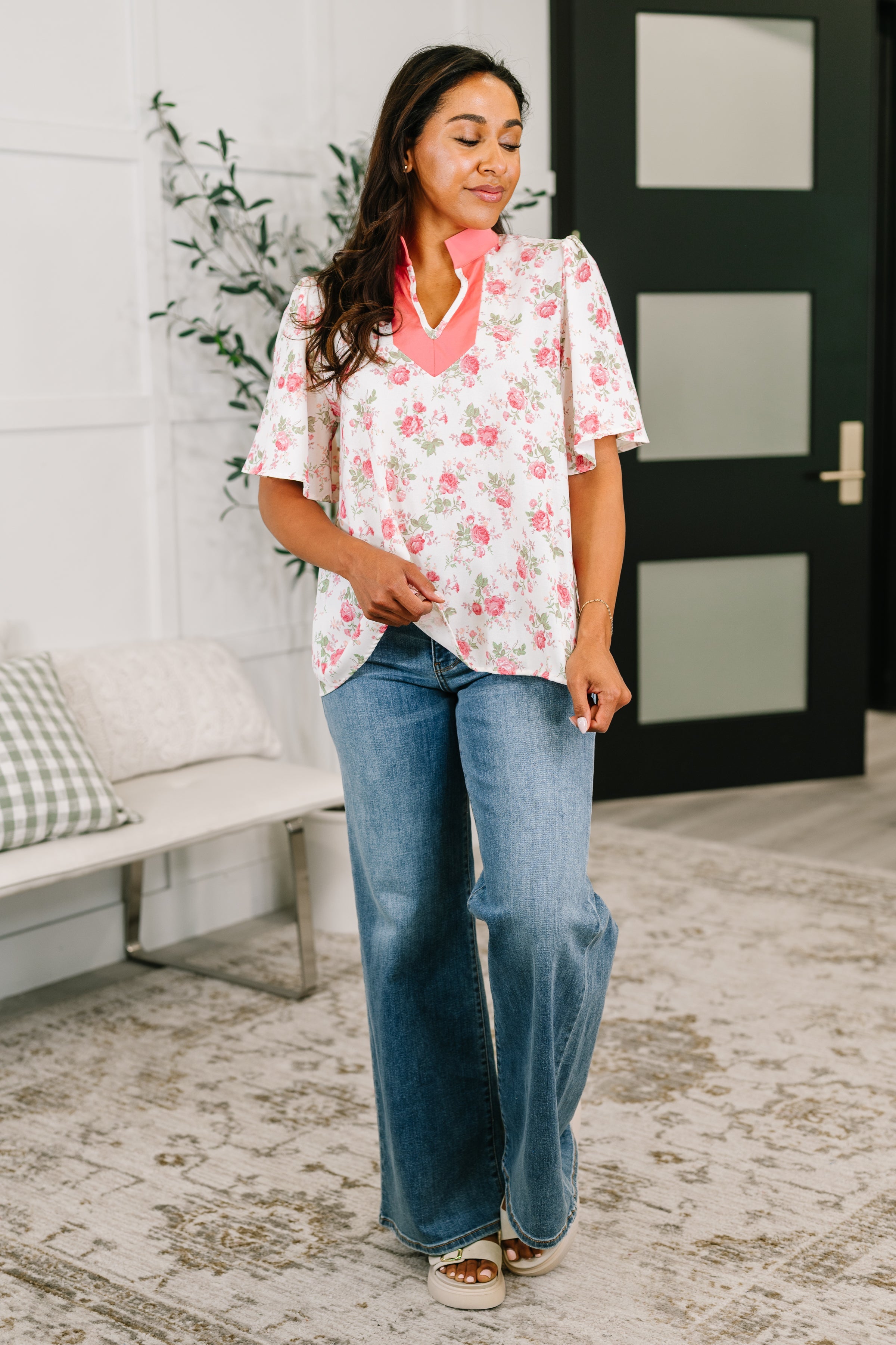 Woman wearing coral-trimmed floral blouse with flutter sleeves paired with wide-leg jeans in eclectic indoor setting