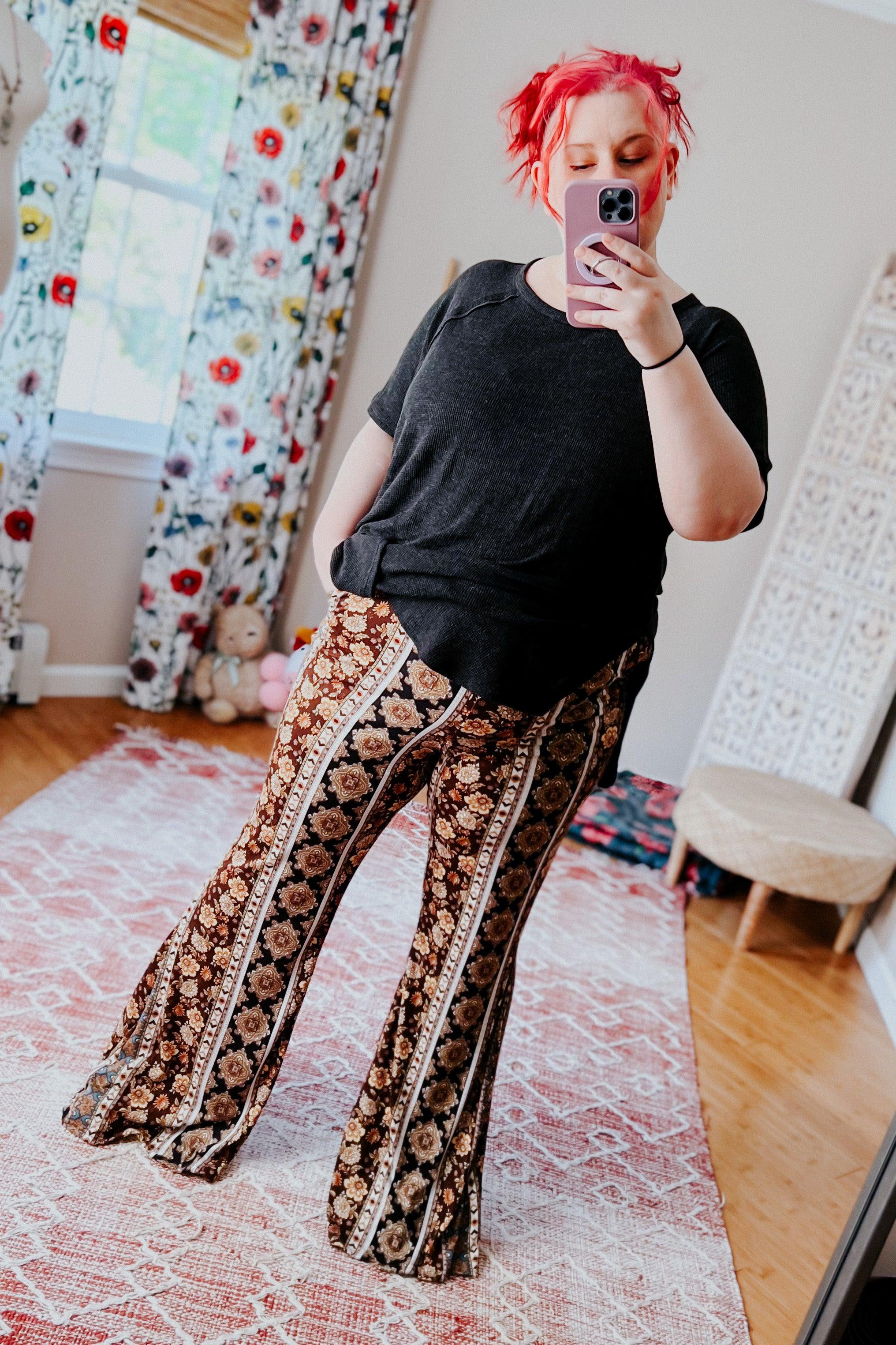 Woman with bright red hair taking mirror selfie wearing black top and floral patterned flared pants in bright room