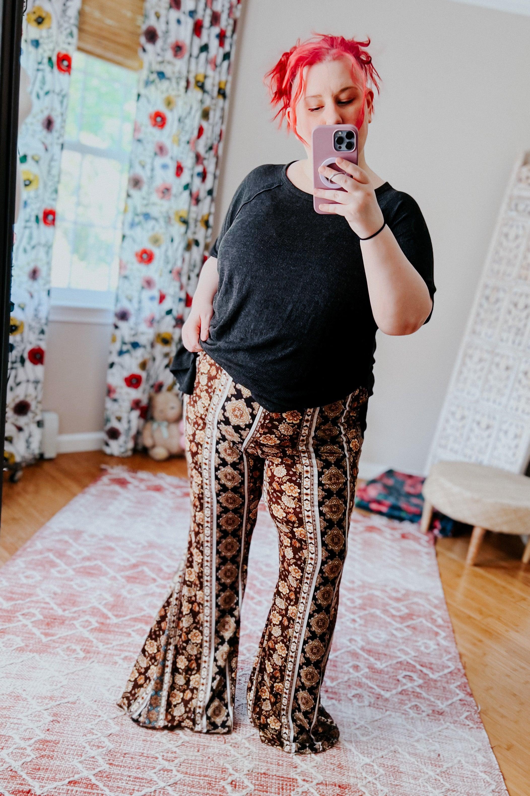 Woman with red hair taking mirror selfie wearing black top and brown floral patterned flare pants in cozy room
