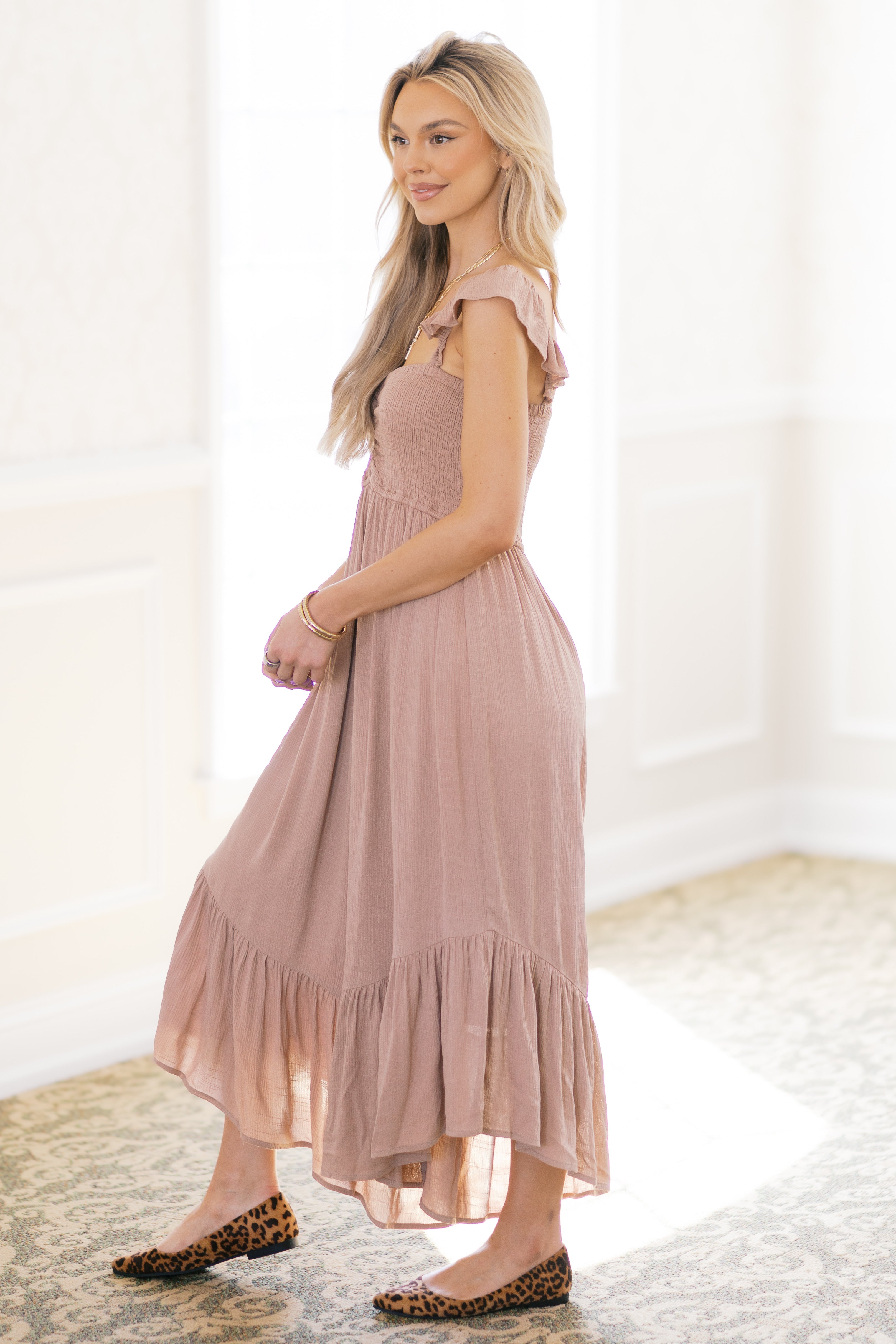 Blonde woman in dusty rose boho midi dress with ruffle sleeves and smocked bodice standing indoors on patterned carpet