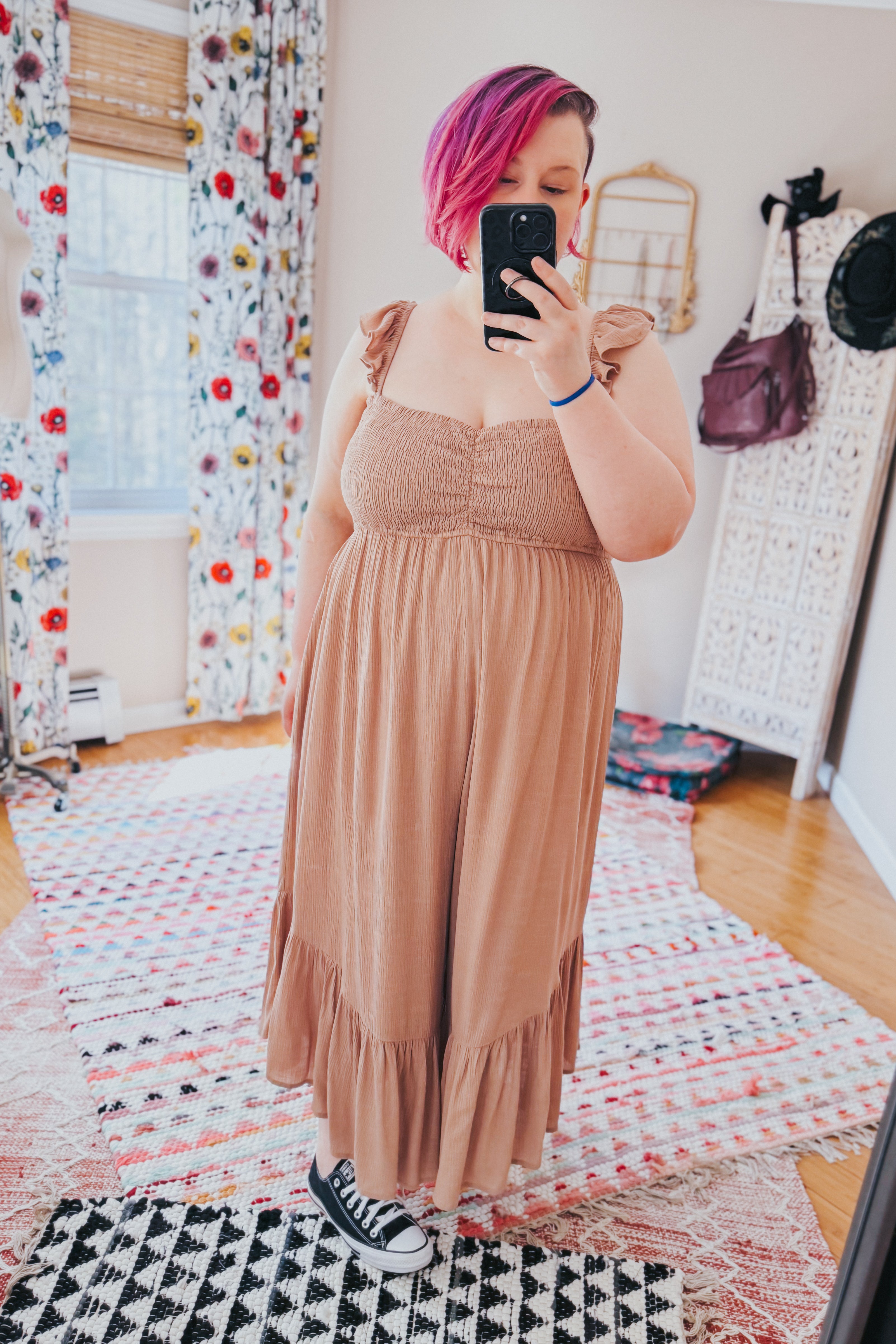 Woman with short pink hair taking mirror selfie wearing light brown ruffle strap wide leg jumpsuit indoors on patterned rugs