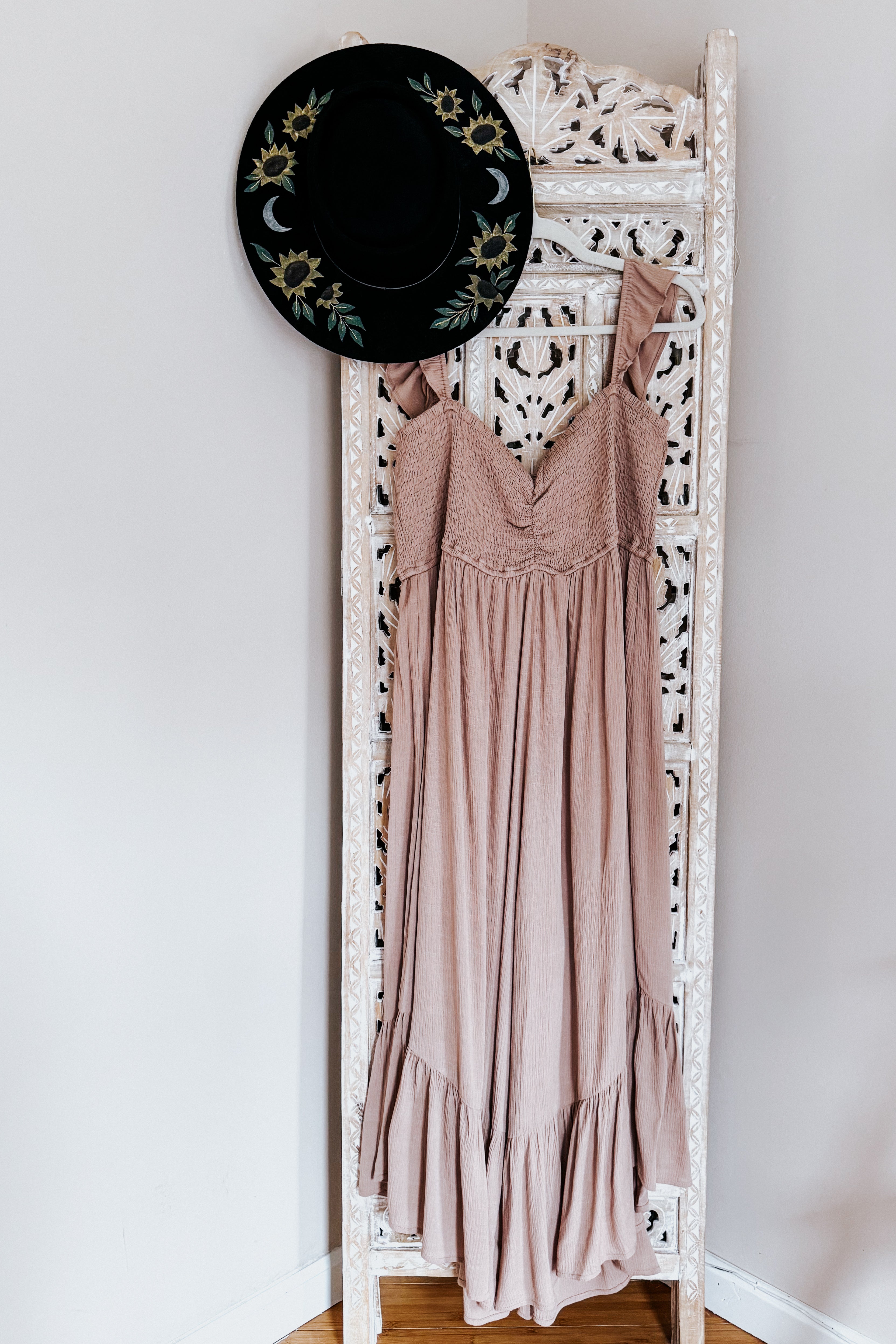 Light brown boho wide leg jumpsuit with ruffle straps hanging on carved white wooden room divider with black floral hat