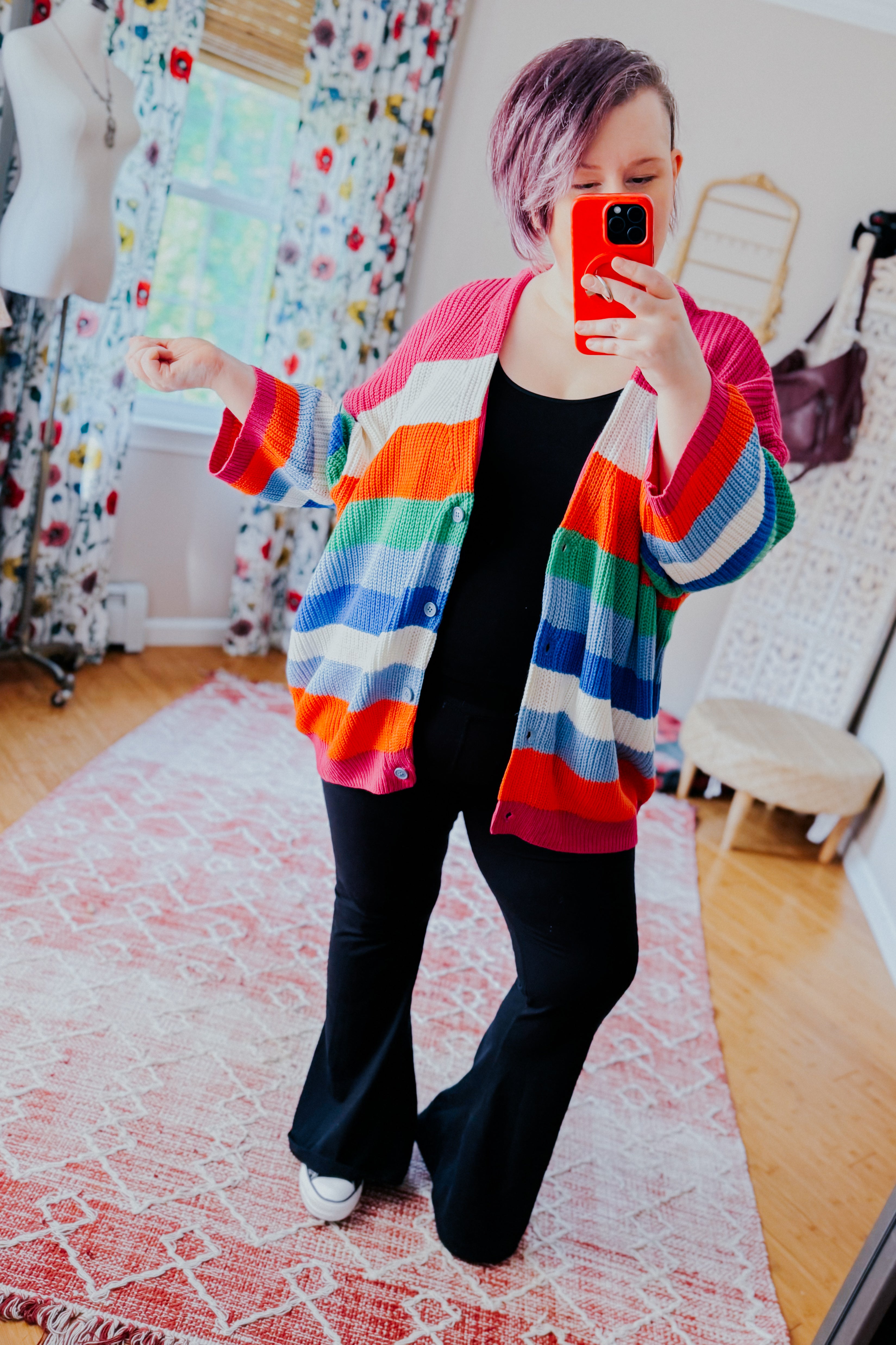 Woman with short hair taking mirror selfie wearing colorful bold striped button down cardigan and black pants indoors