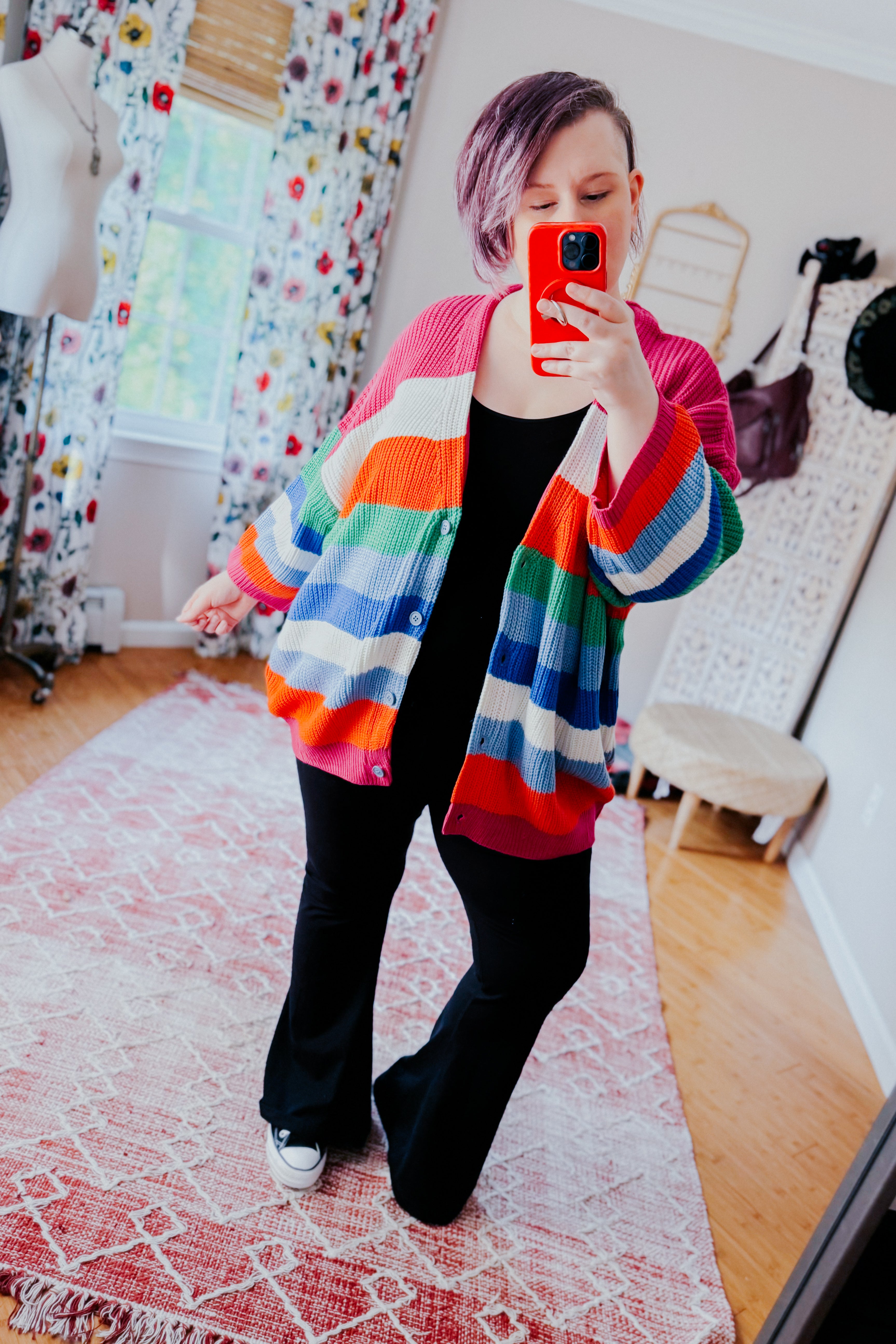 Woman taking mirror selfie wearing colorful bold striped button down cardigan with black flared pants indoors