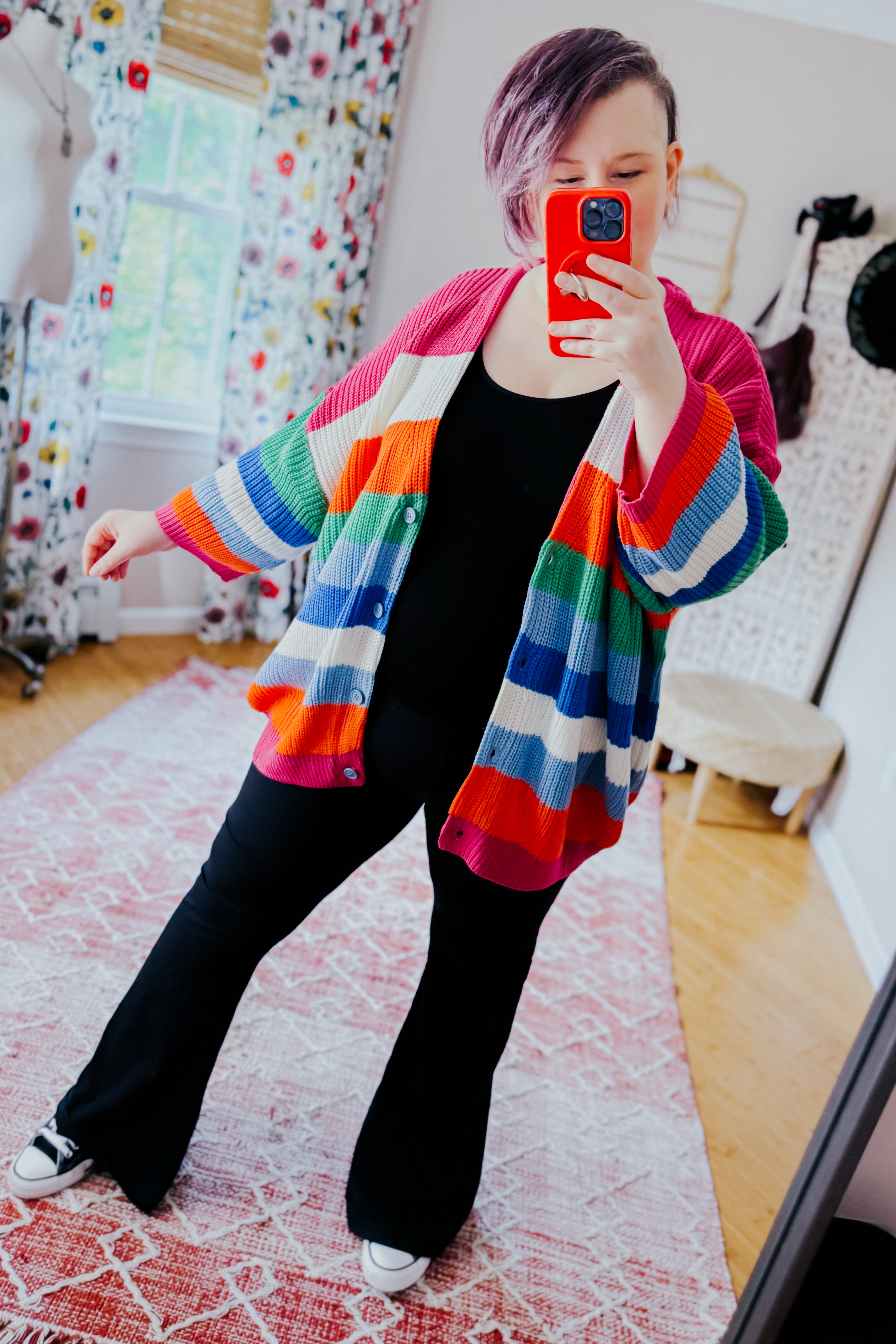 Woman taking mirror selfie wearing colorful striped button down cardigan with black top and pants indoors