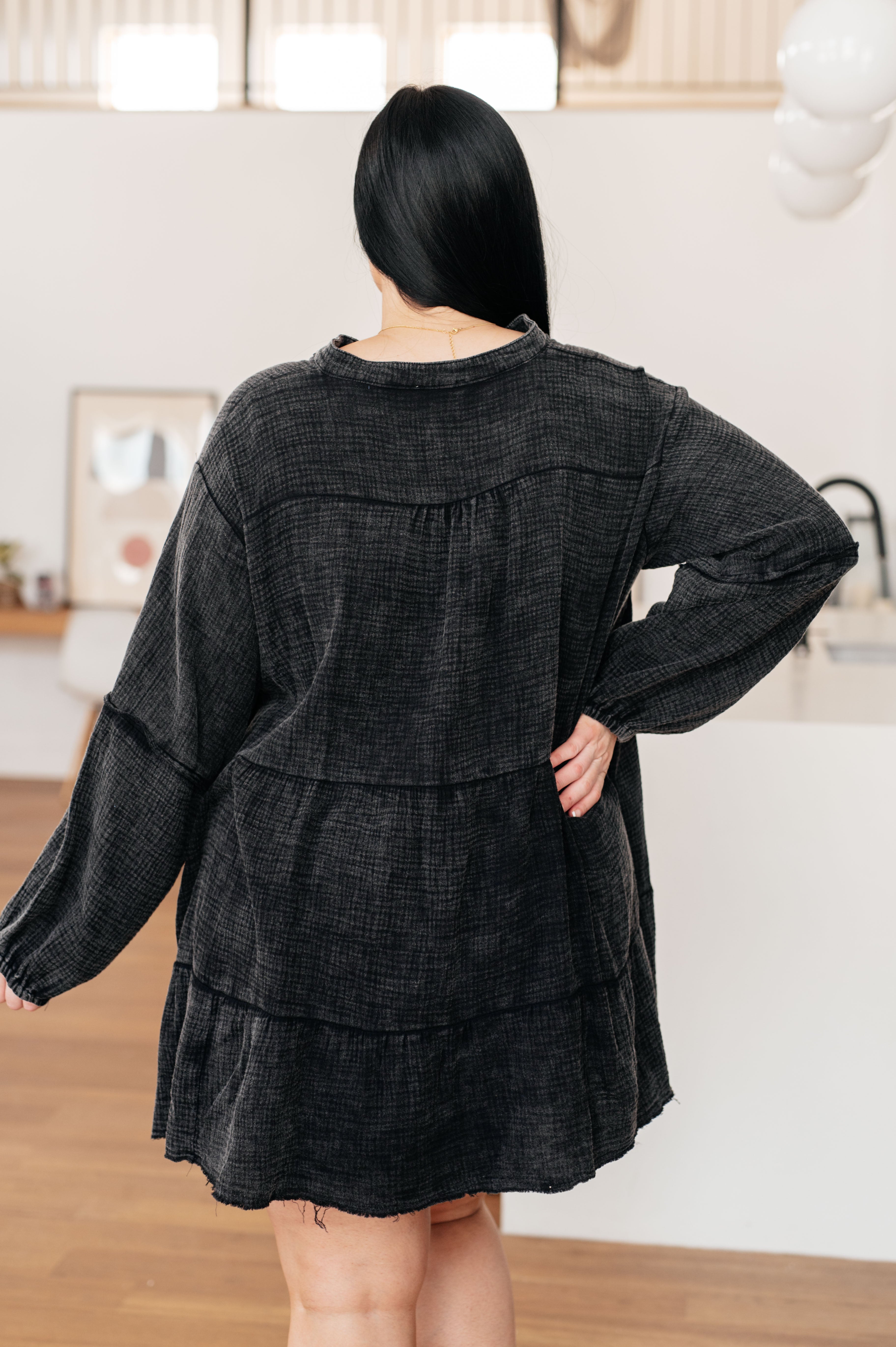 Woman with long dark hair wearing loose black mineral wash dress with tiered back detail standing indoors