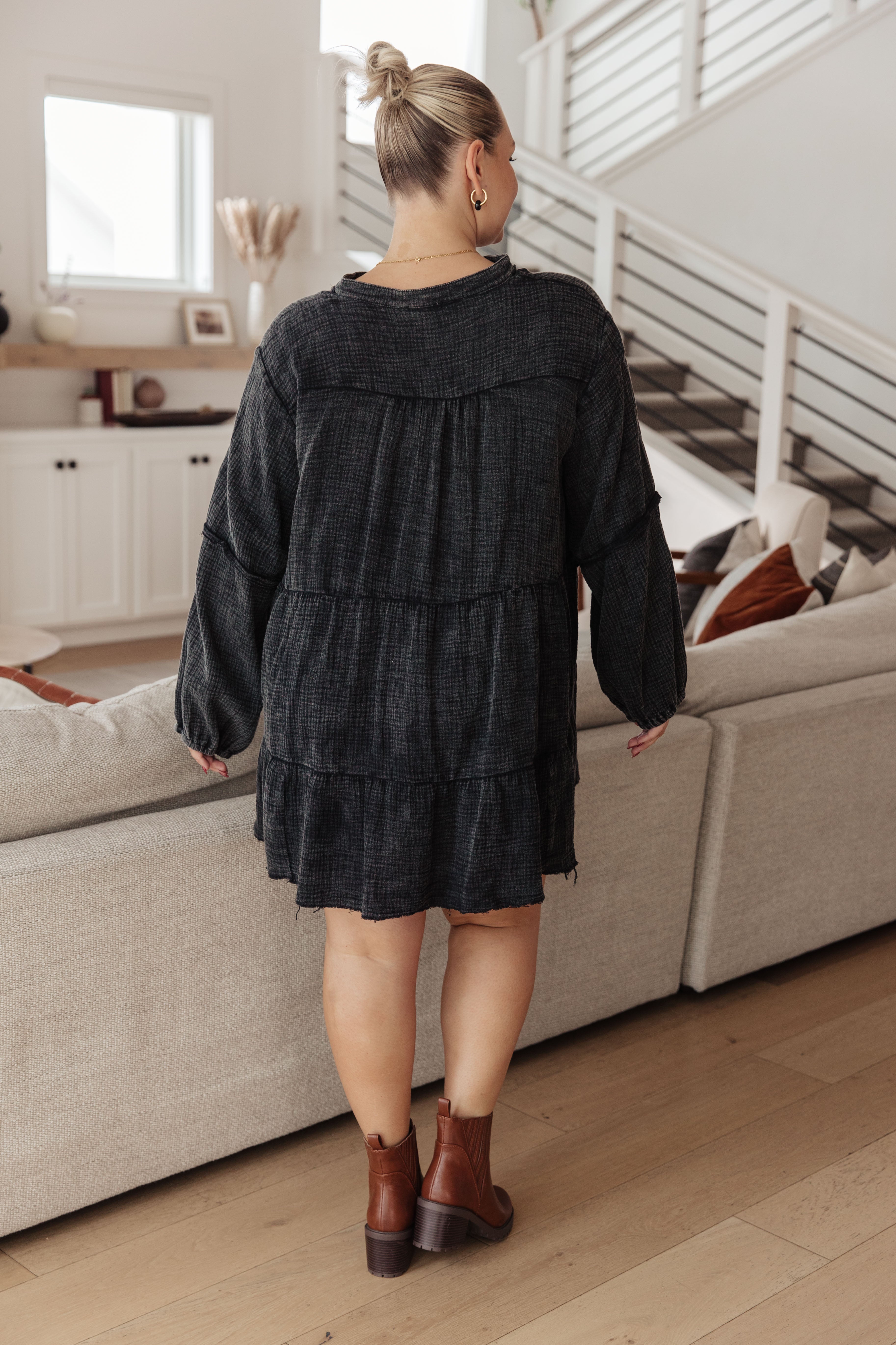 Woman wearing loose dark mineral wash tiered dress with long sleeves and brown ankle boots standing indoors
