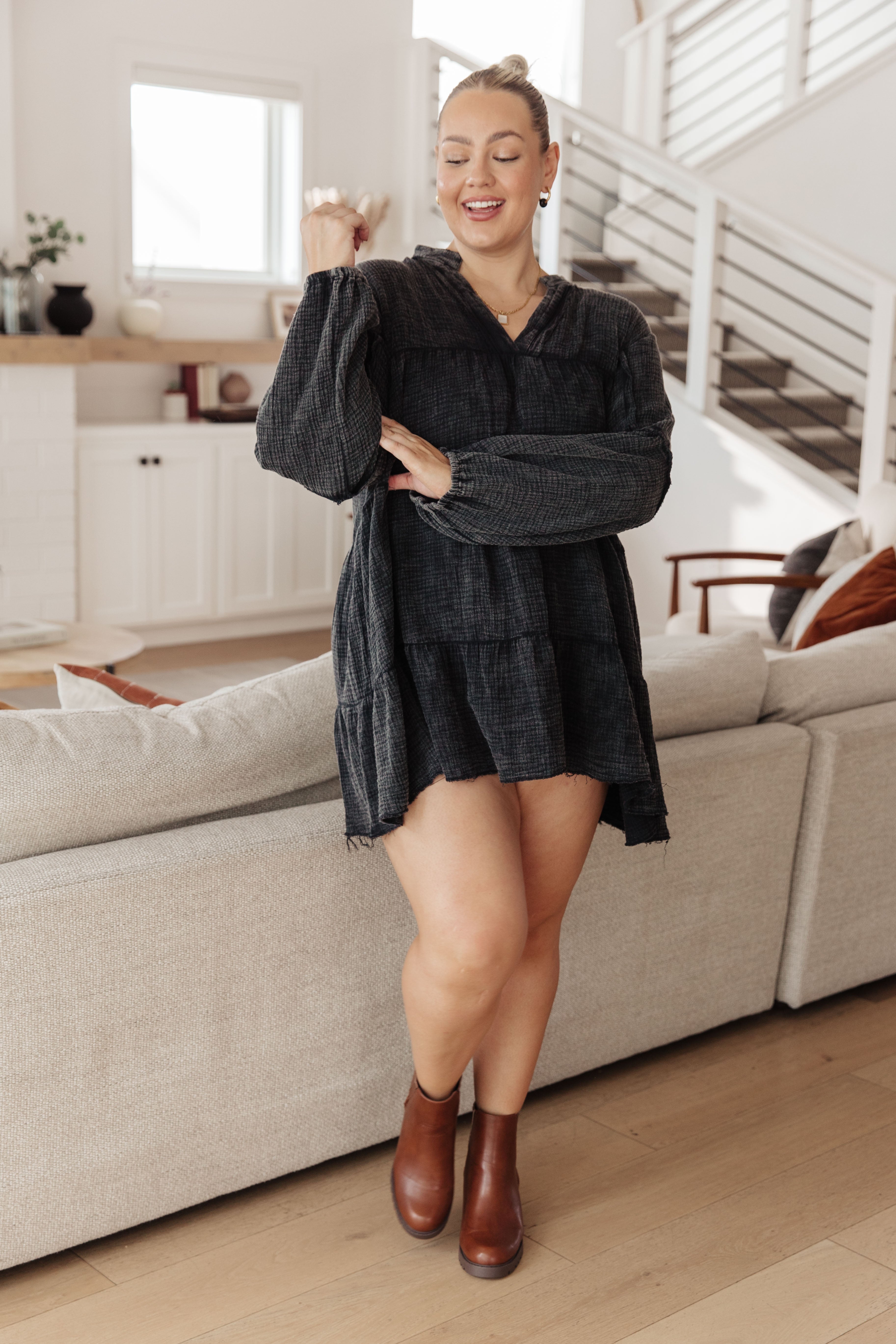 Smiling woman in loose dark mineral wash V-neck dress with balloon sleeves and brown ankle boots standing in modern living room