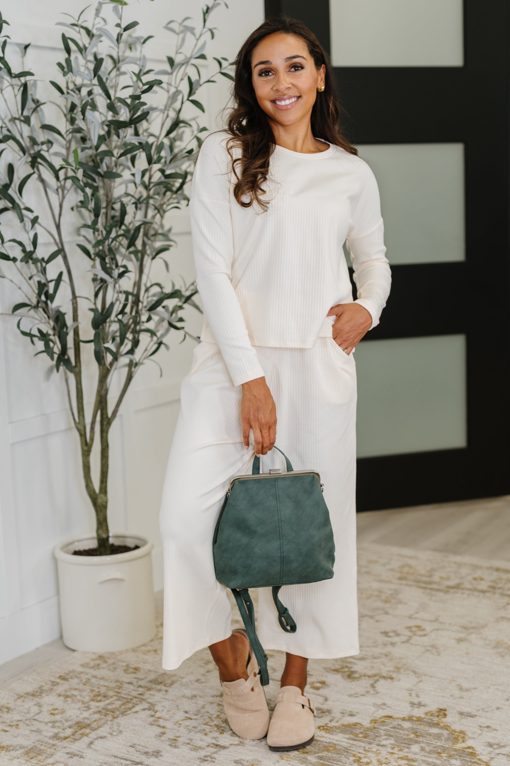 Smiling woman in cream loungewear holding a green dainty backpack with floral lining in bright, eclectic room