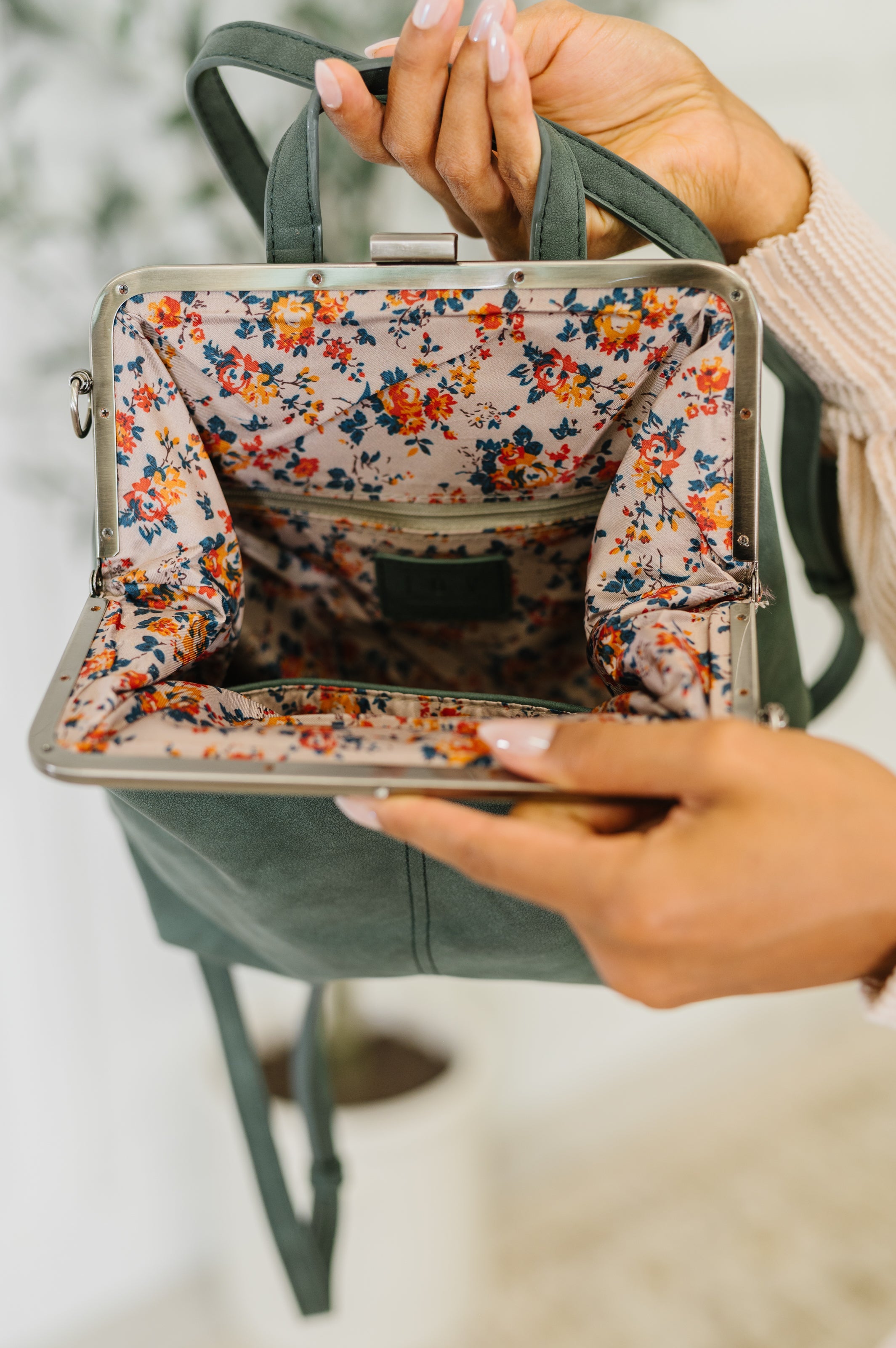 Hands holding open green backpack showing floral print lining and metal frame clasp closure inside