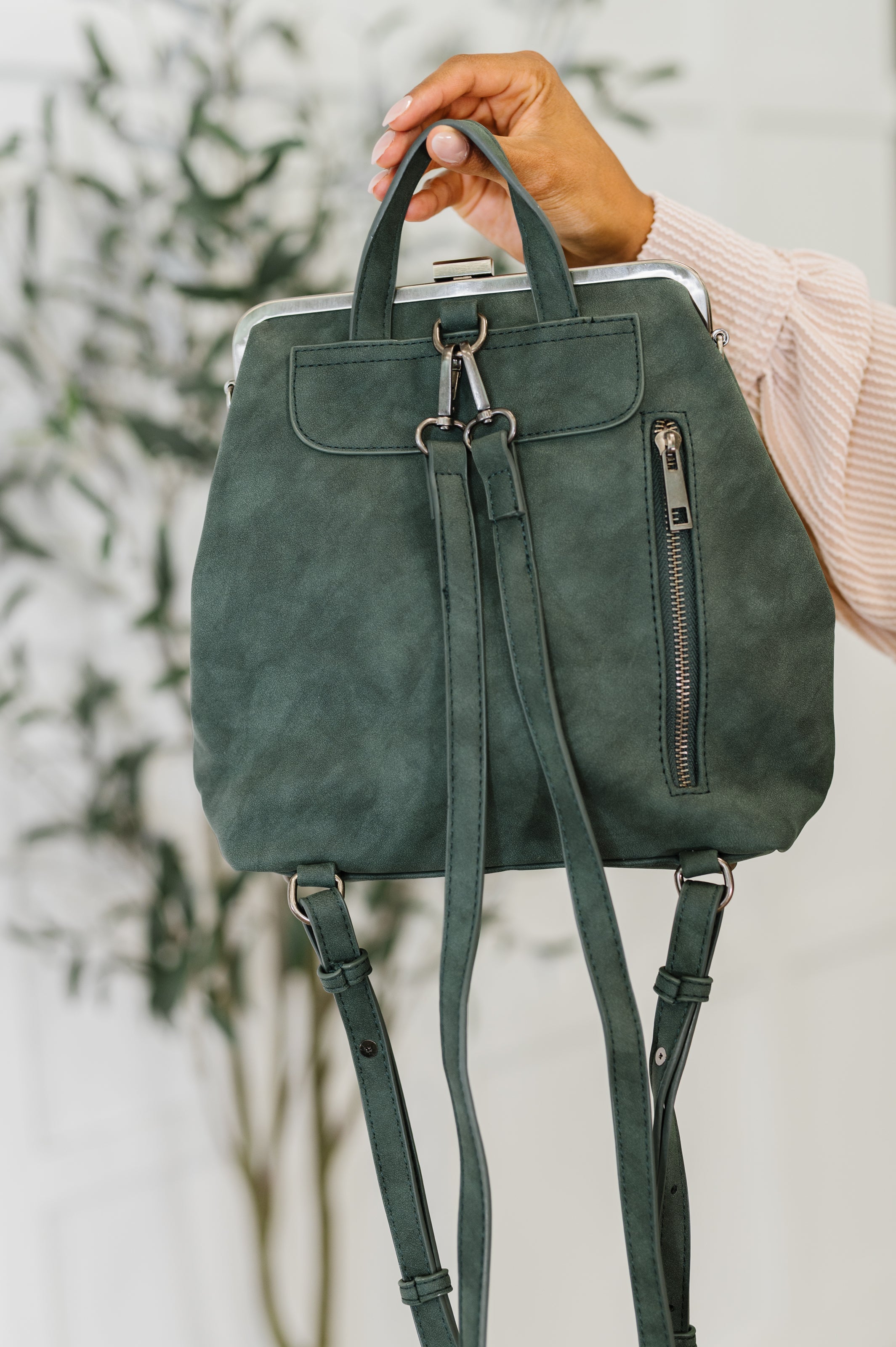 Hand holding green suede backpack with silver zipper and adjustable straps against blurred indoor plant background