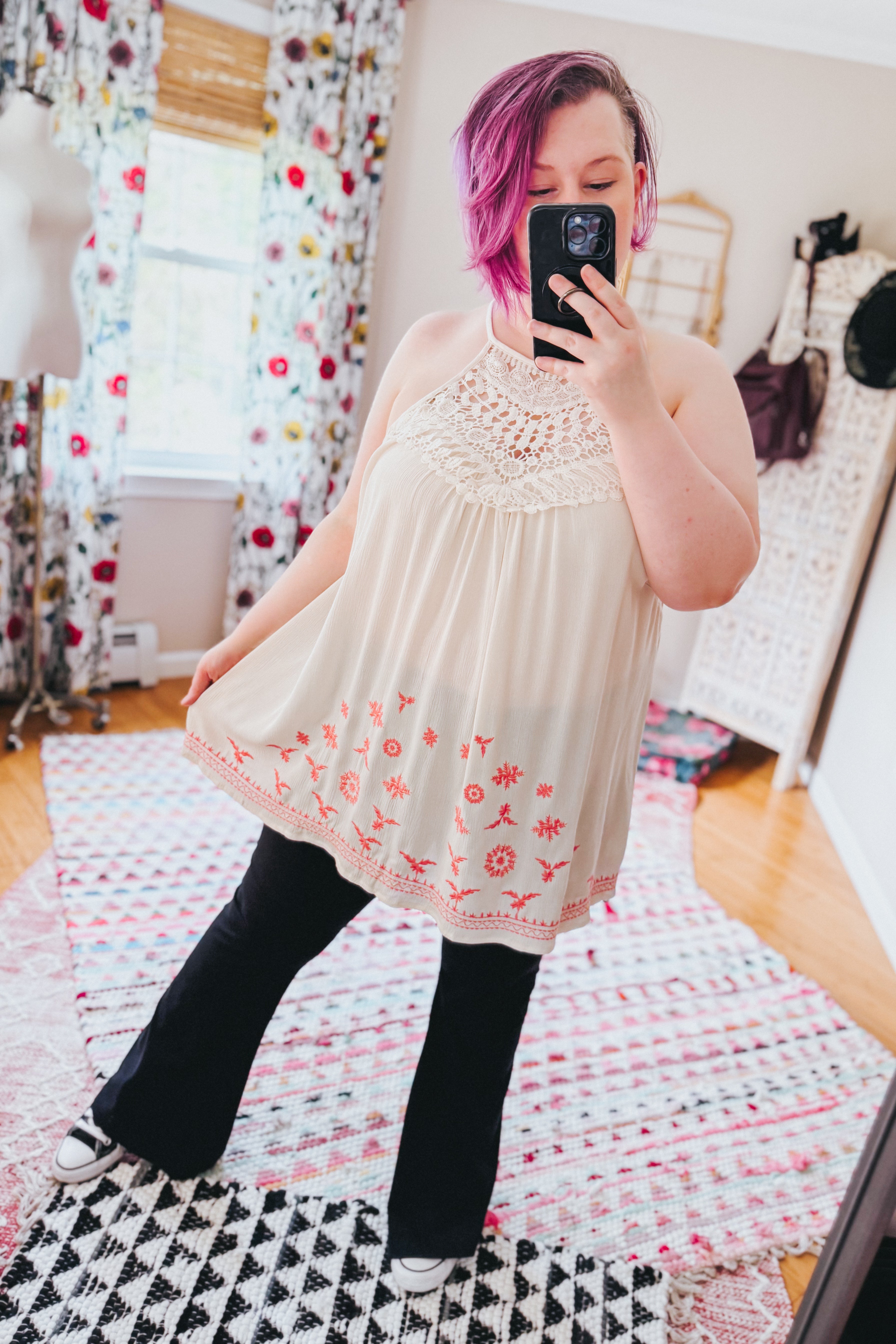 Woman wearing cream halter top with crochet neckline and orange floral embroidery paired with black pants indoors