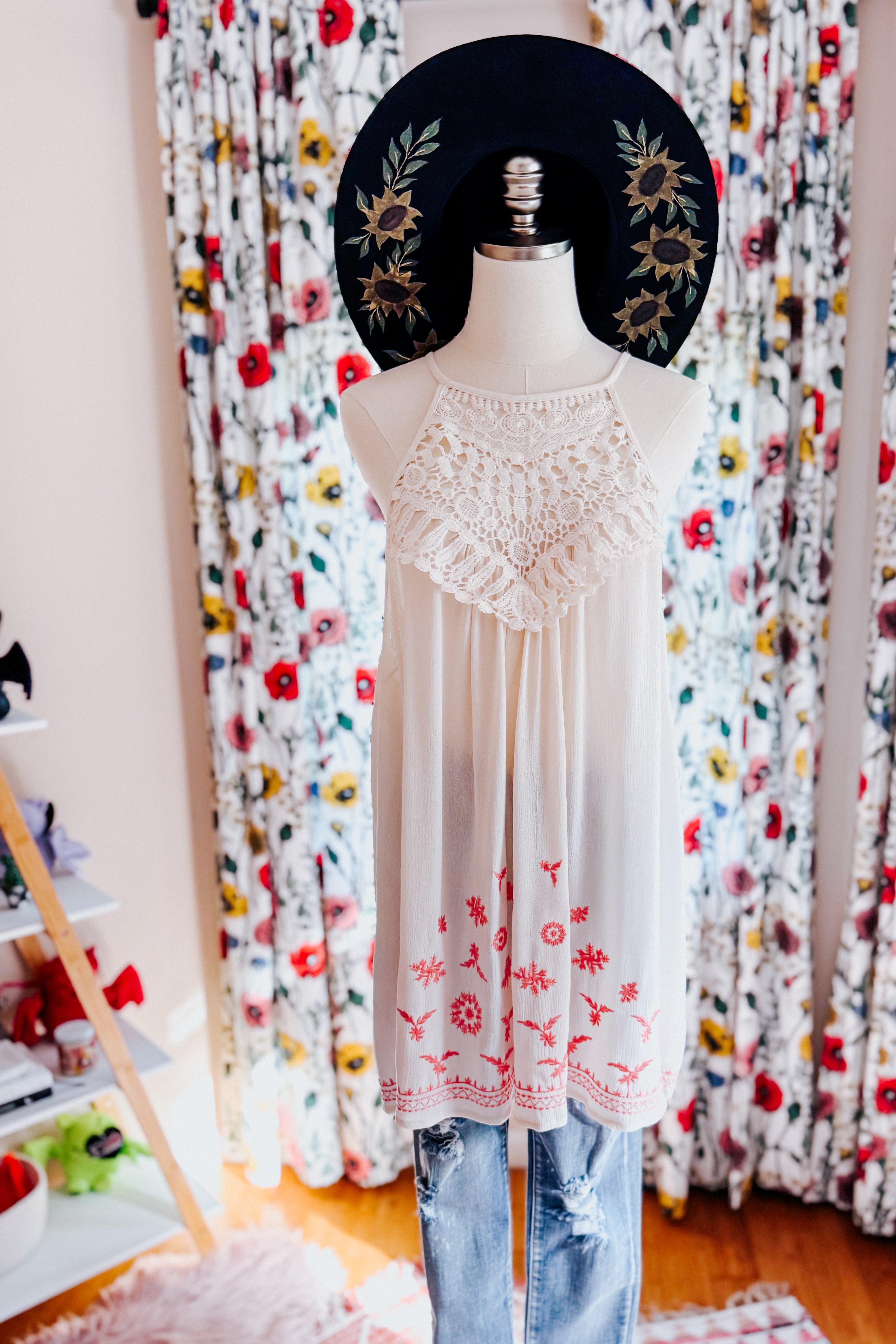 Cream sleeveless top with crochet detailing and red floral embroidery paired with distressed blue jeans on mannequin indoors