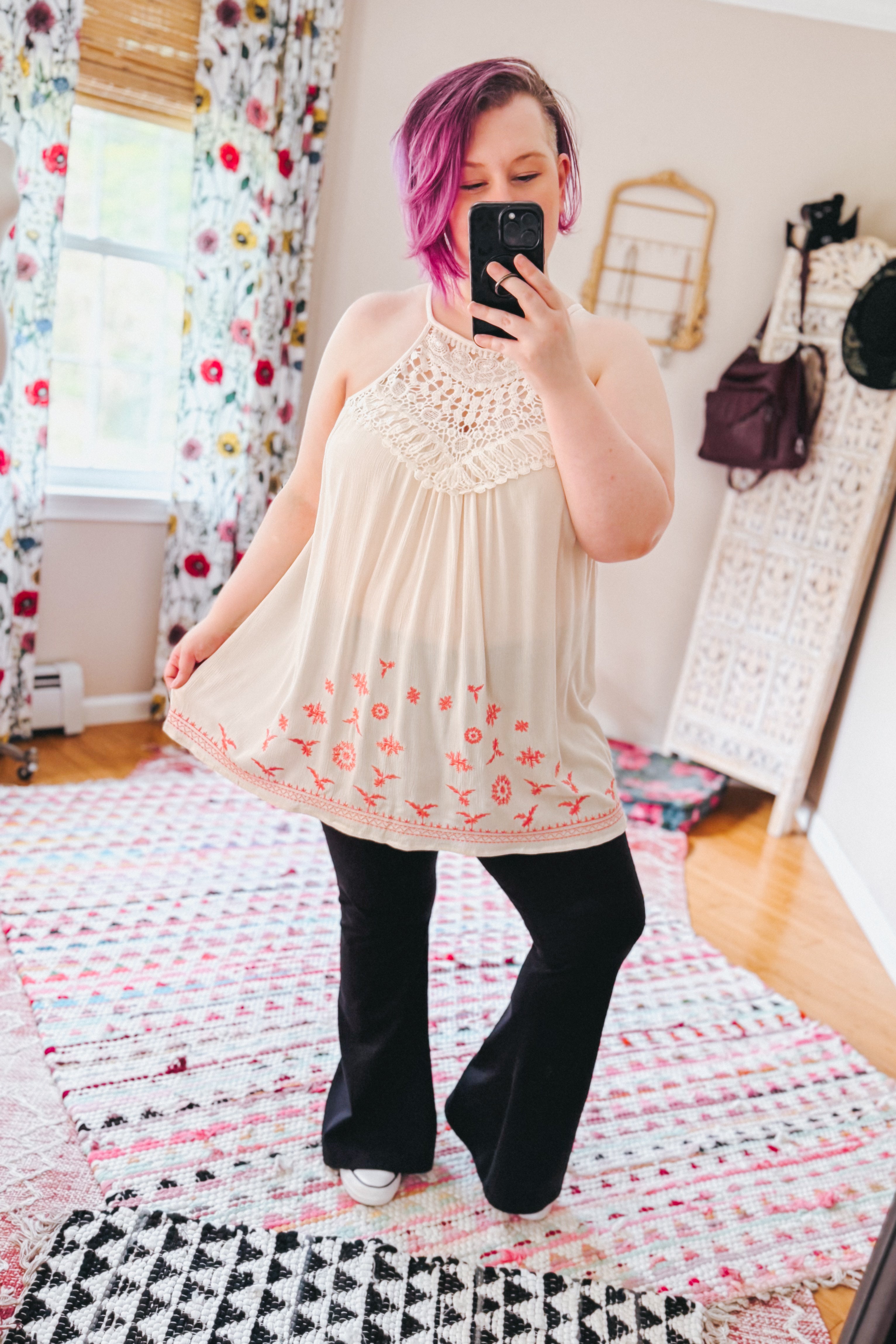Woman wearing a cream halter neck top with crochet detail and coral floral embroidery paired with black flared pants indoors