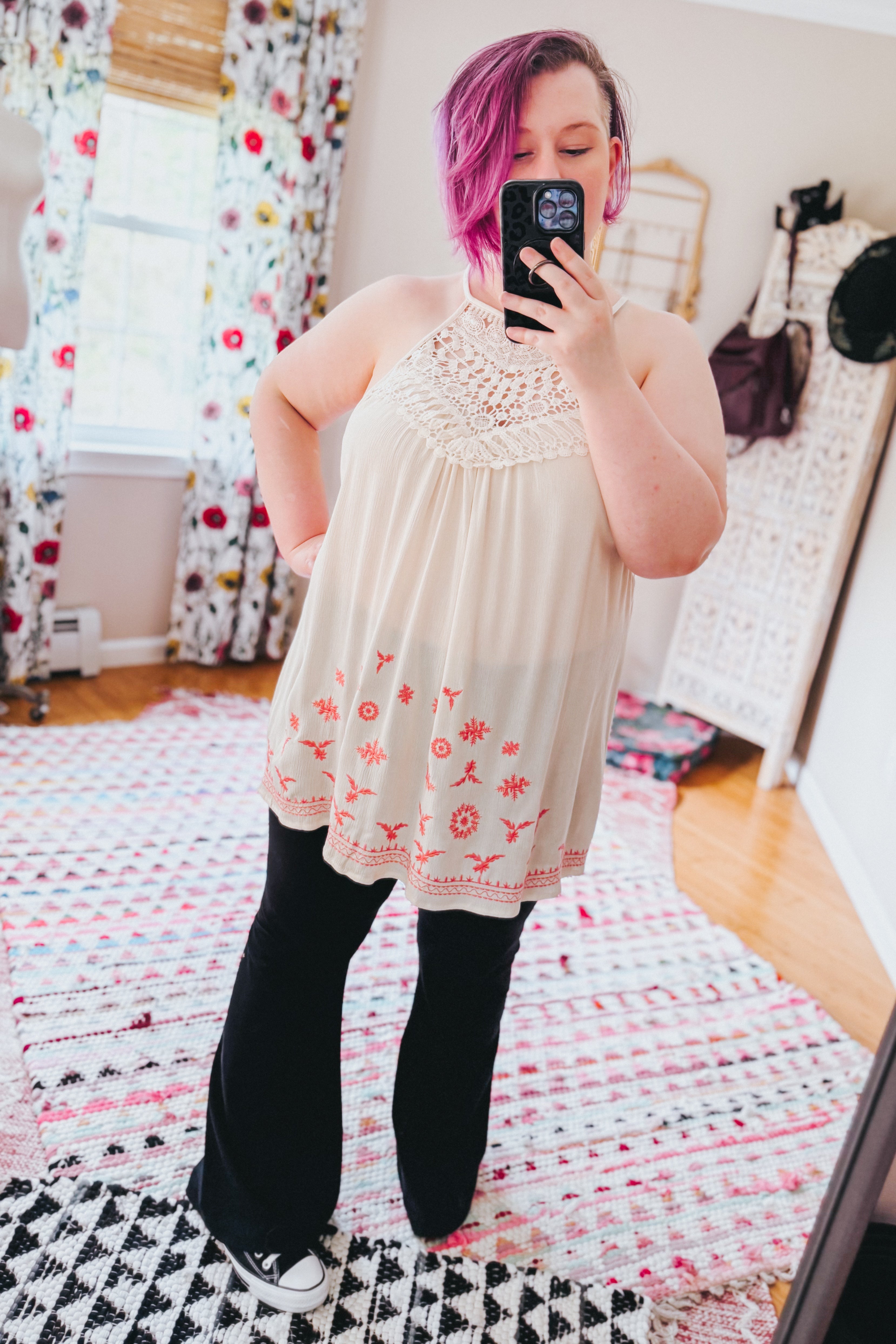 Woman taking mirror selfie wearing cream sleeveless top with crochet yoke and red floral embroidery paired with black pants indoors