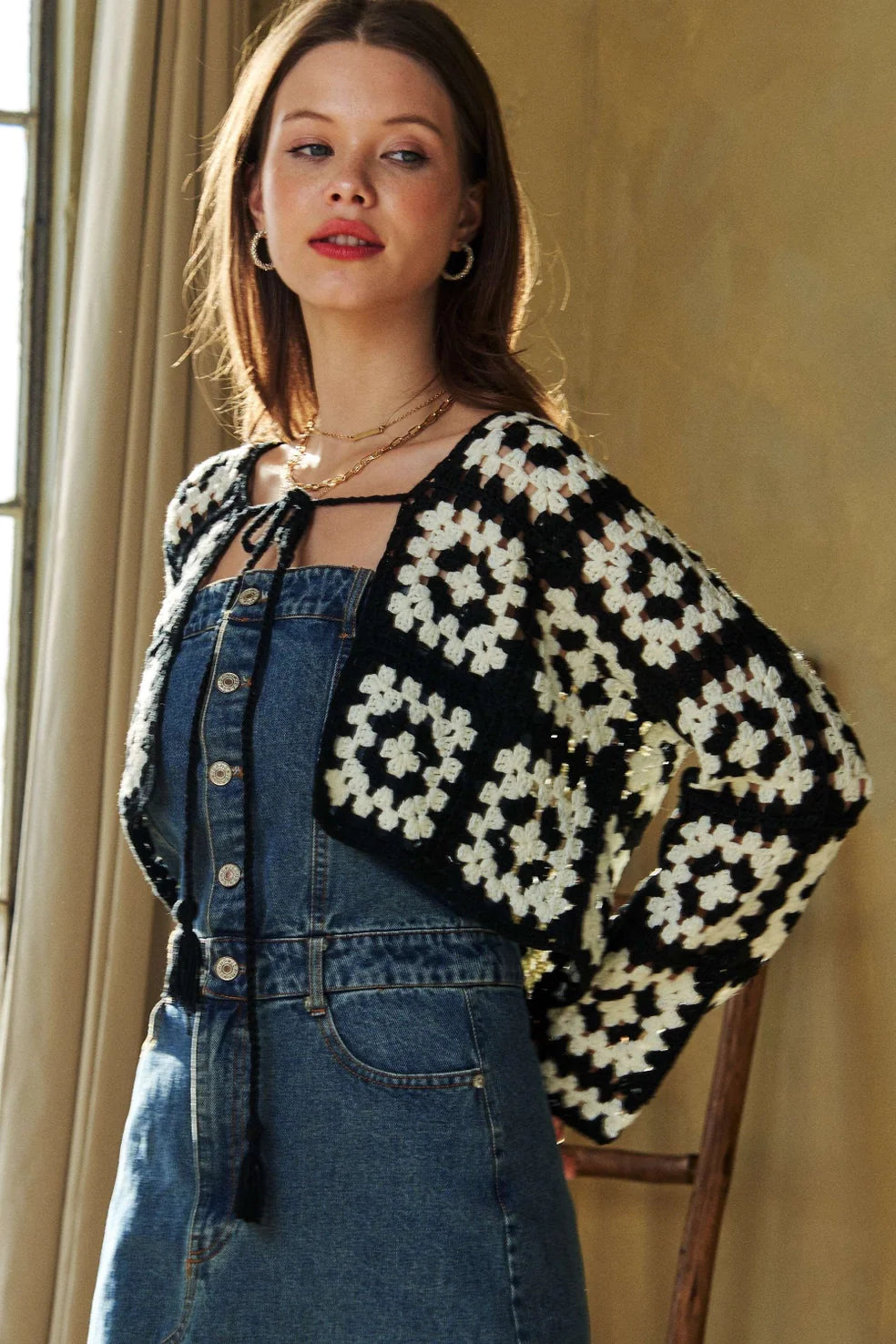Model indoors wearing black and cream crochet cropped cardigan over a button-up denim dress with layered gold necklaces