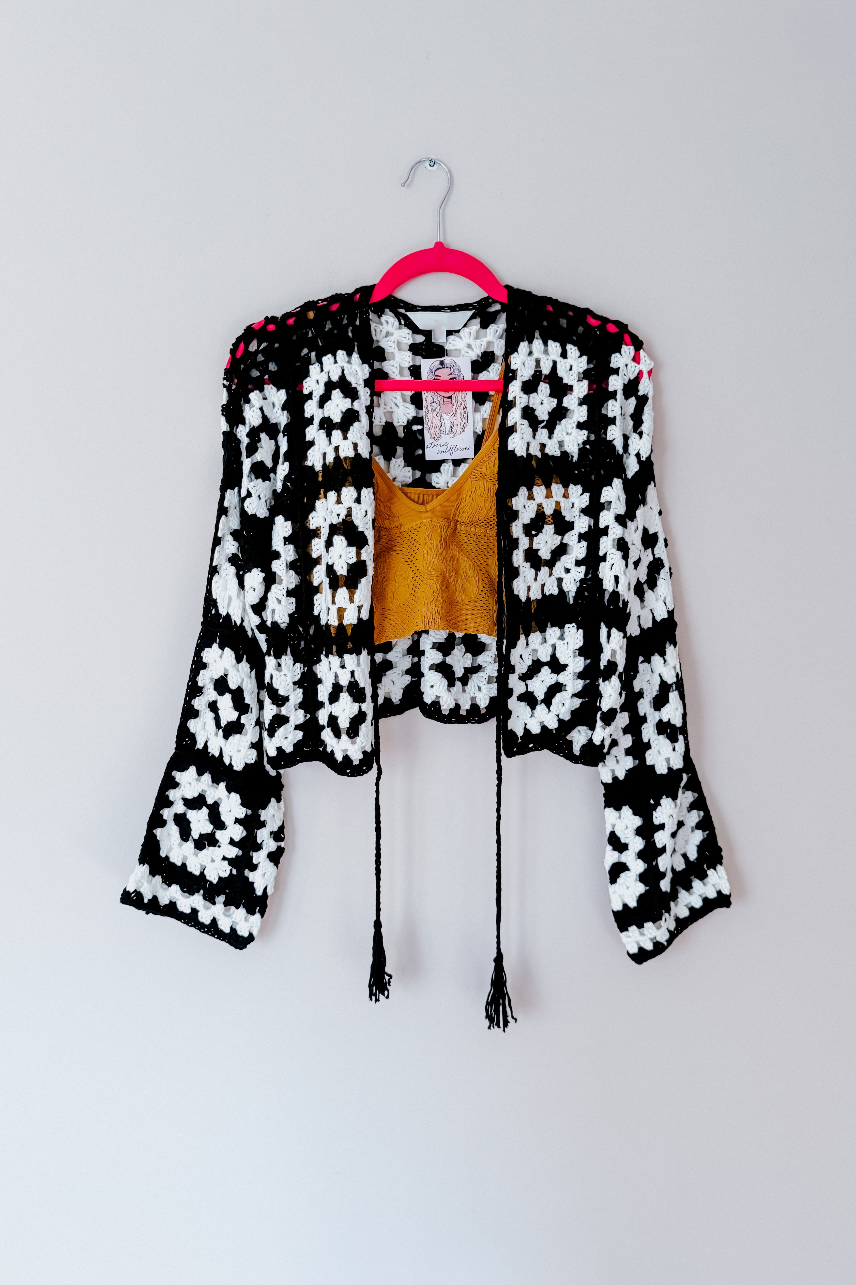 Black and white lightweight crochet cropped cardigan with long sleeves hanging on pink hanger against white background