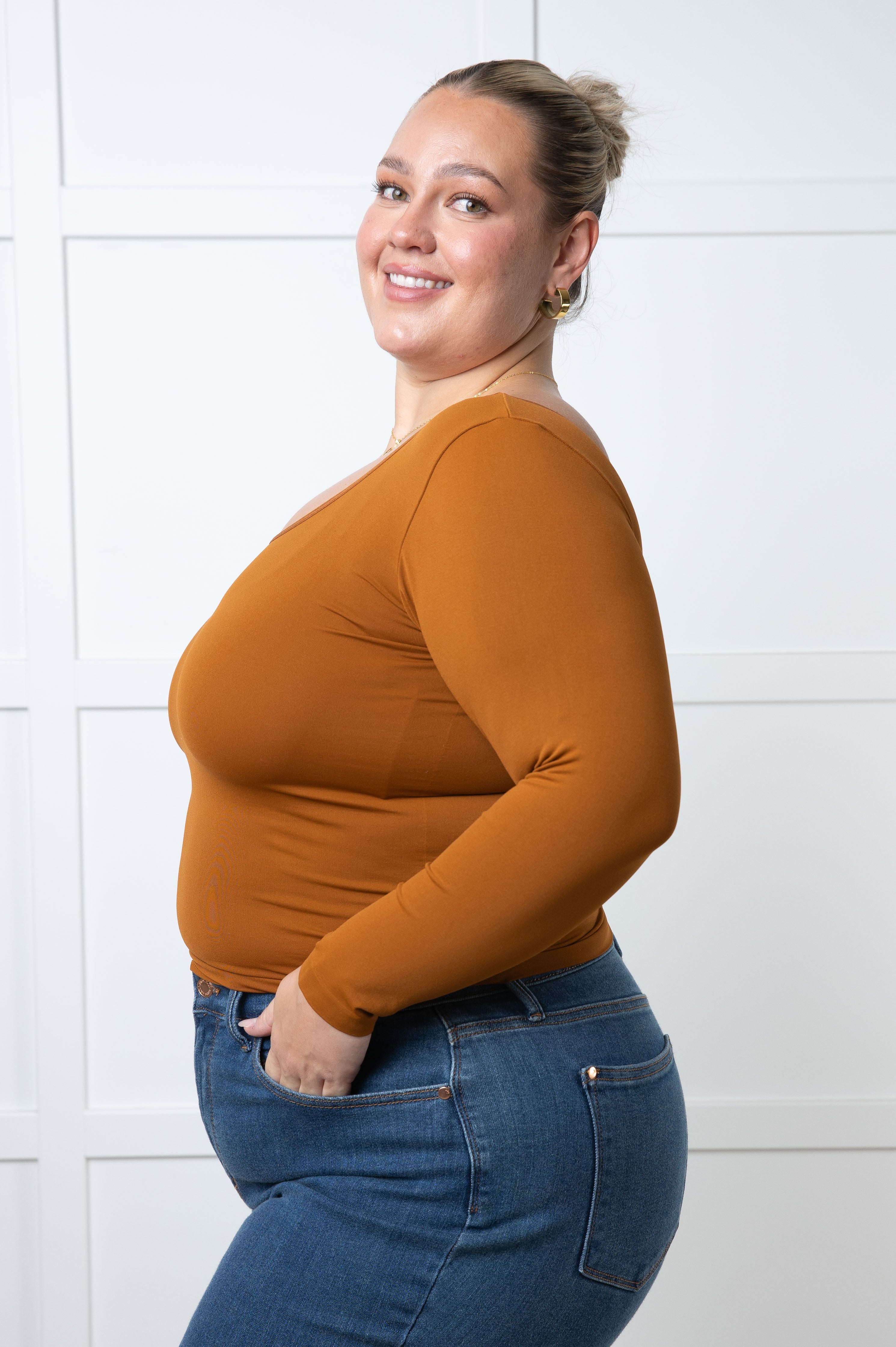Smiling woman wearing caramel seamless V-neck long sleeve top with blue denim jeans against white background