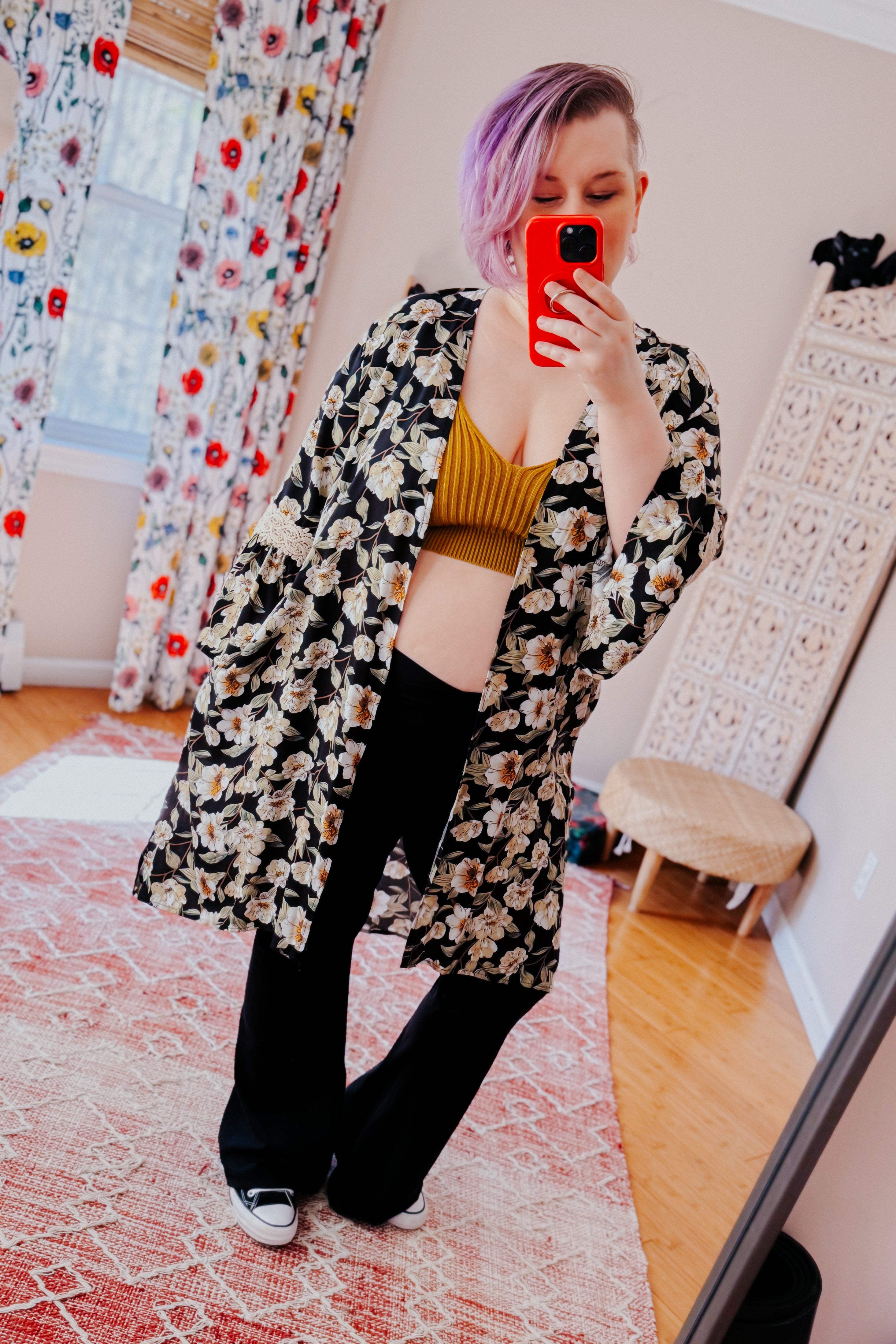 Woman with short purple hair wearing black floral 3/4 bell sleeve duster over mustard crop top and black pants taking selfie indoors