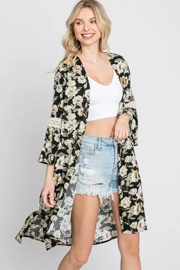 Blonde woman wearing black floral print 3/4 bell sleeve duster with lace trim over white crop top and ripped denim shorts